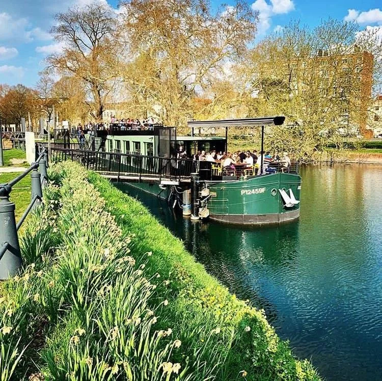 Une péniche avec des passagers se trouvant sur une rivière, près d’un parc avec des arbres feuillus et des fleurs au bord de l’eau, sous un ciel bleu avec quelques nuages.