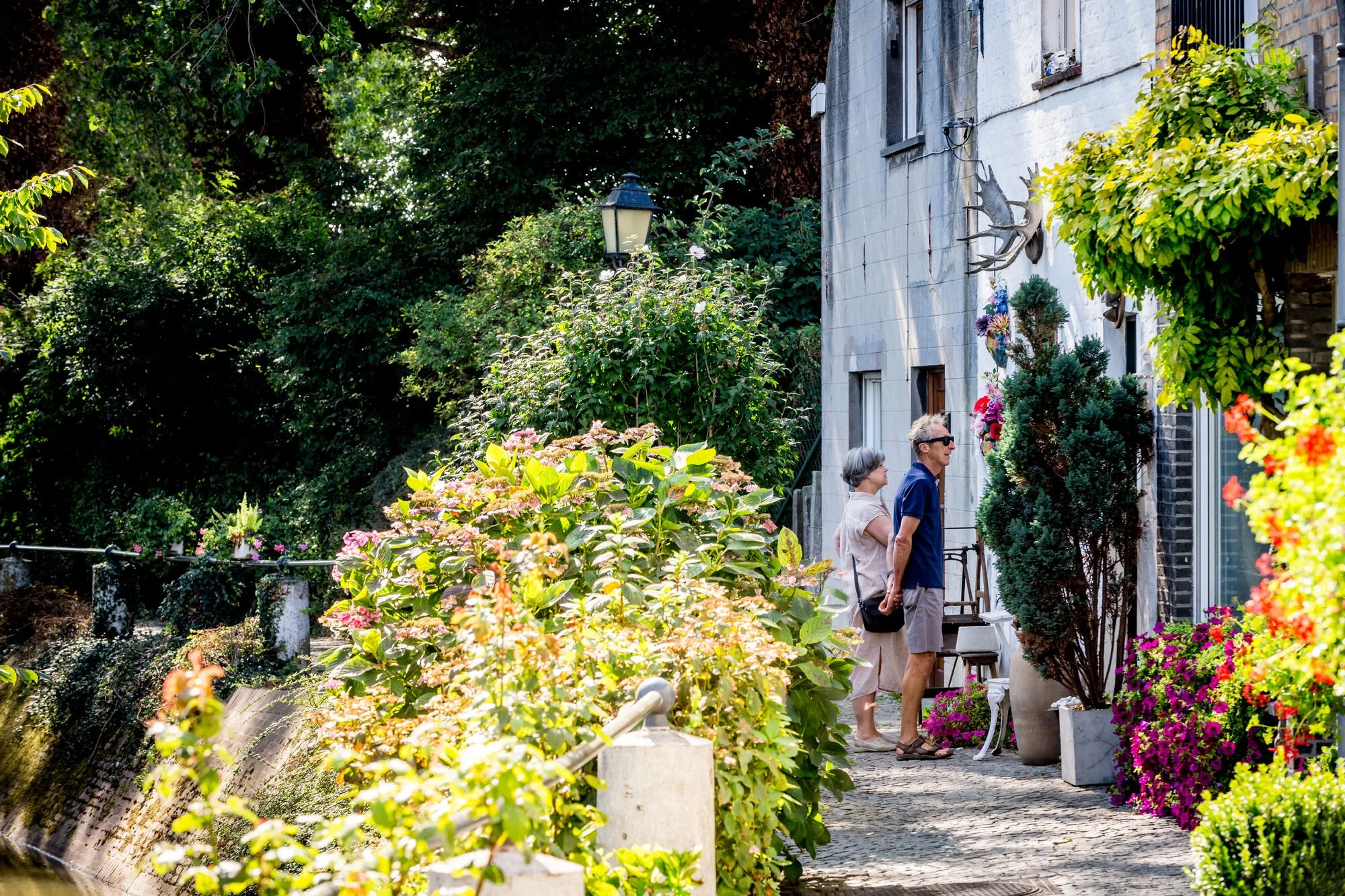 Deux personnes âgées, un homme portant des lunettes de soleil et une femme, regardent devant une maison ornée de fleurs et de décorations de chasseurs, avec un jardin verdoyant et un lampadaire visible.