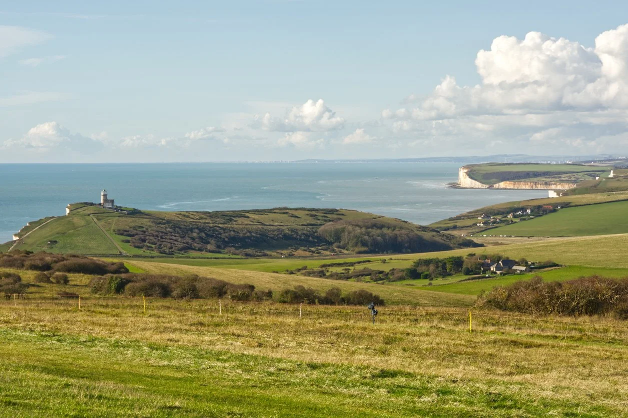 Paysage côtier avec falaises, un phare en haut d'une colline verte, et la mer en arrière-plan, sous un ciel partiellement nuageux.