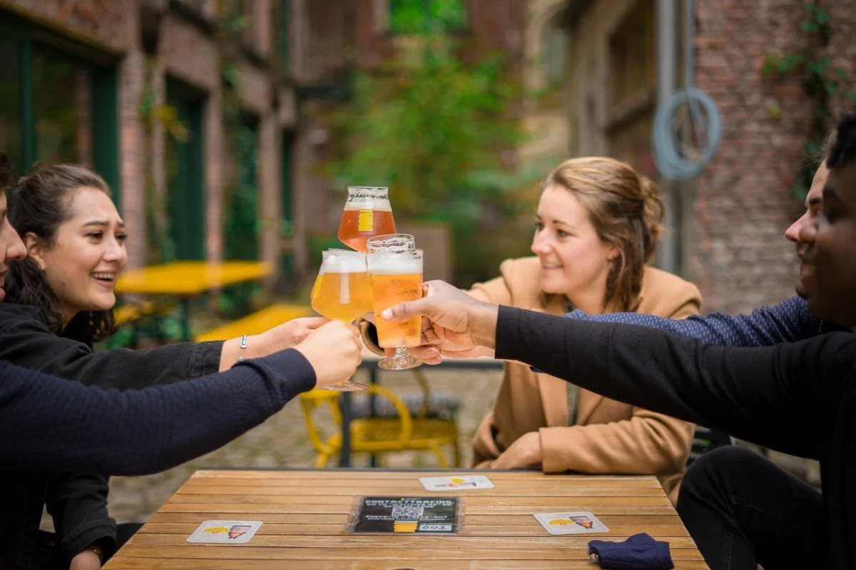 Quatre personnes souriantes trinquent avec des verres de bière dans une cour extérieure en pierre, avec des bancs et des tables en bois. La scène est joyeuse et conviviale.