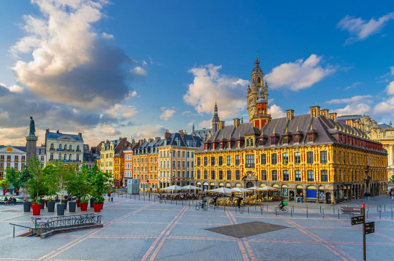 Une place urbaine avec des bâtiments colorés, une fontaine, des arbres en pot, des petites personnes et des cyclistes, sous un ciel partiellement nuageux.