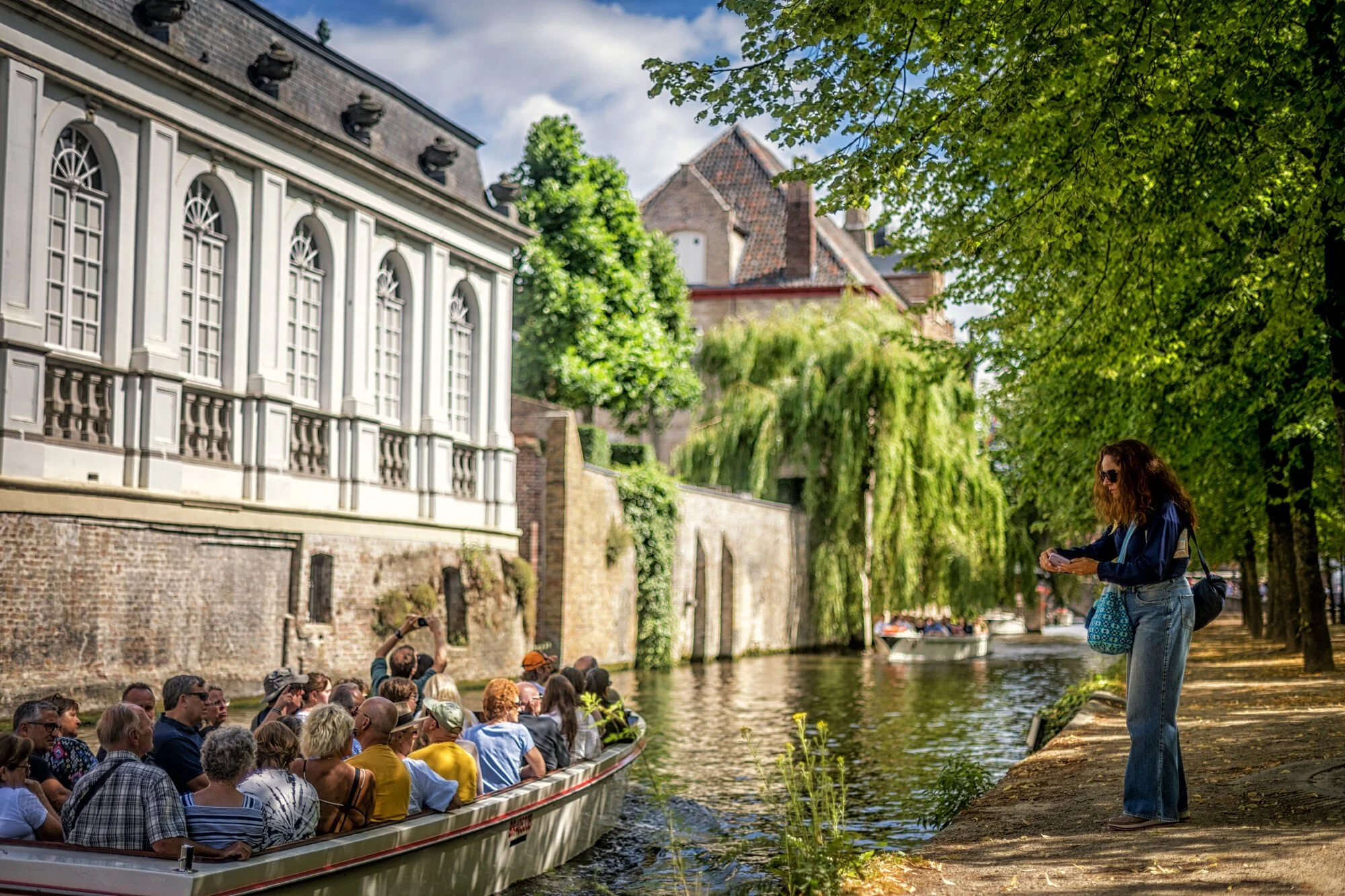 Une femme avec des cheveux bouclés, portant des lunettes de soleil, un sweat à capuche et un jean, en train de lire ou regarder quelque chose dans ses mains en se tenant sur le trottoir près d'une allée bordée d'arbres feuilles vertes, côté canal où passe une barque remplie de touristes. La scène se déroule dans un endroit pittoresque avec des bâtiments historiques et un ciel partiellement nuageux.