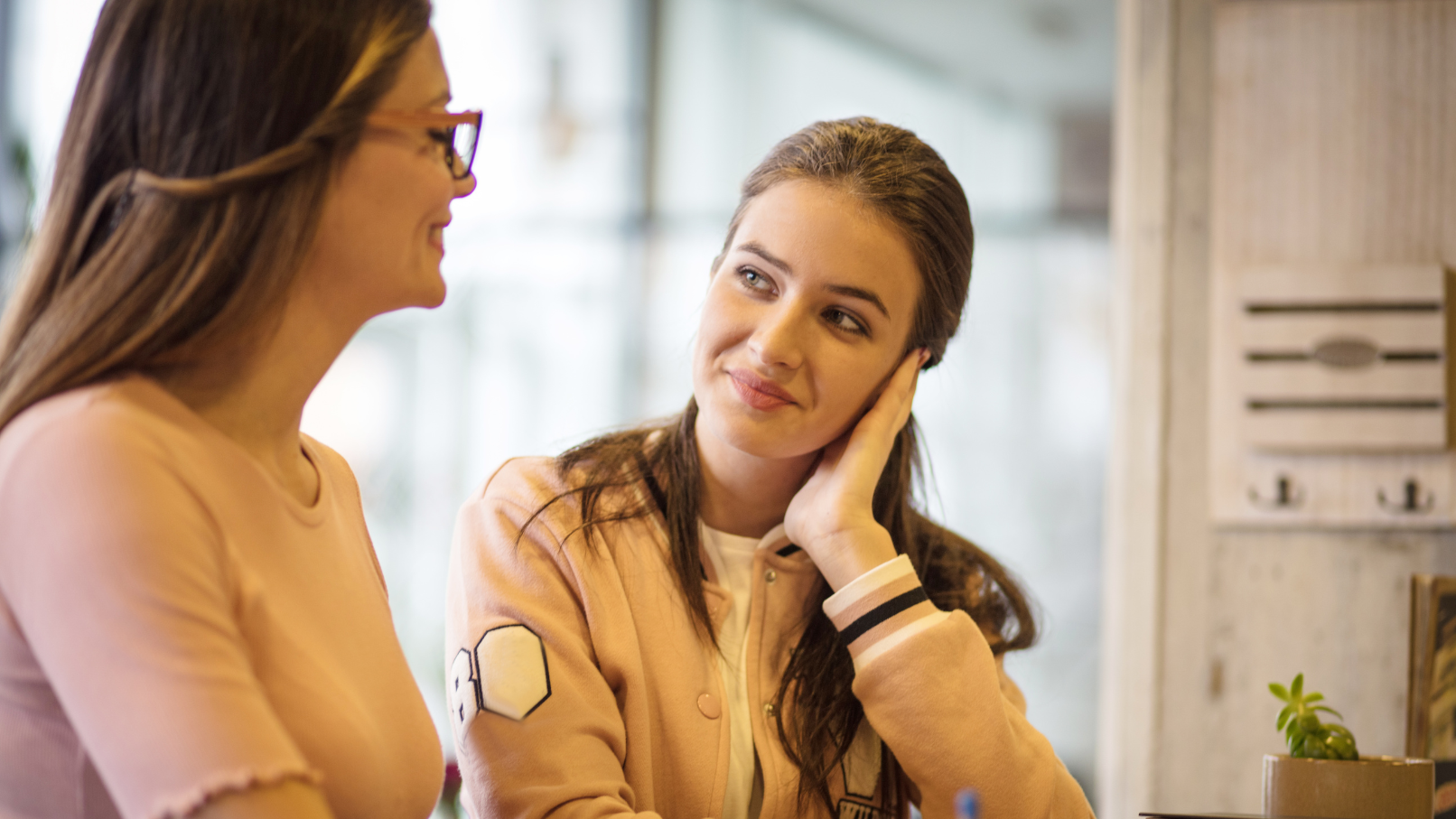 Two young women sitting at a table, engaging in a friendly conversation, one smiling and the other listening. They are indoors with natural light.