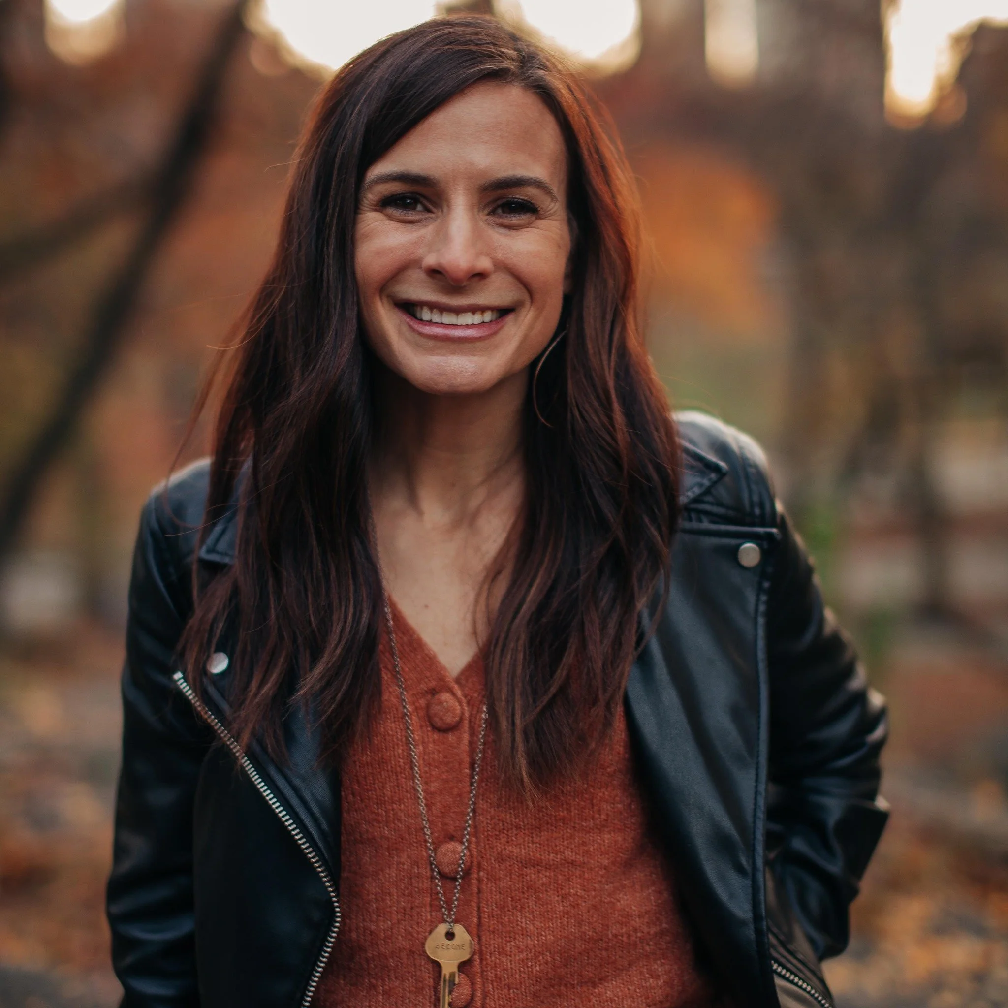 A woman with long dark hair smiling outdoors in autumn with trees and fallen leaves in the background.