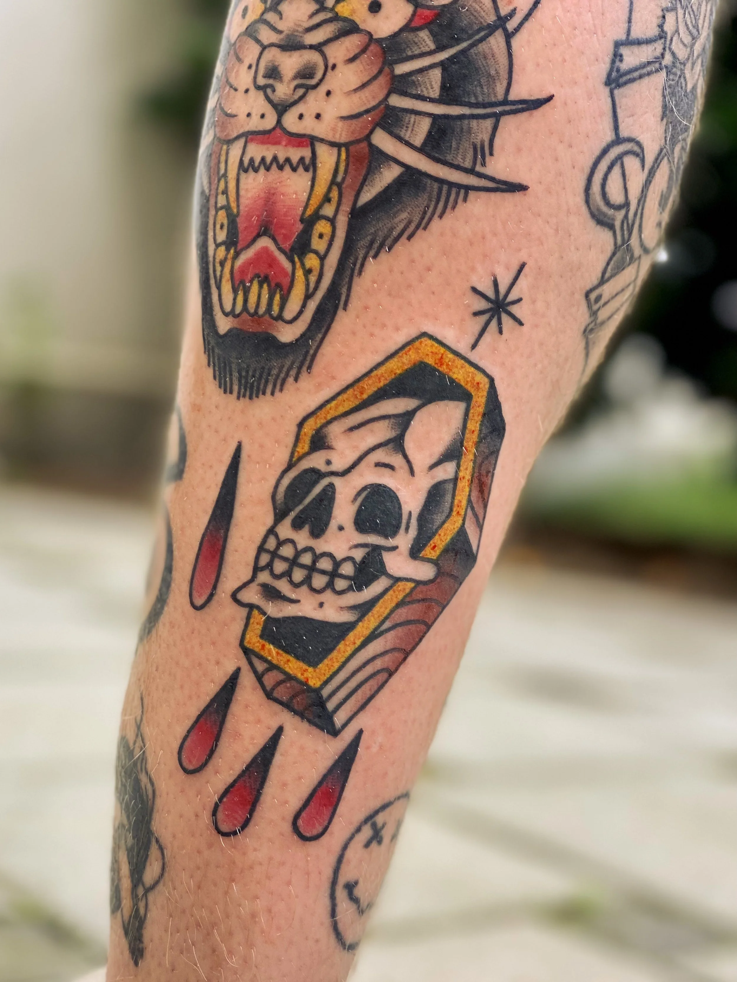 Tatovering af en åben kranie med store øjenhuler og tænder, omgivet af dråber, og en tiger med åbent fanget munden, en sort stjerne og andre geometriske elementer.