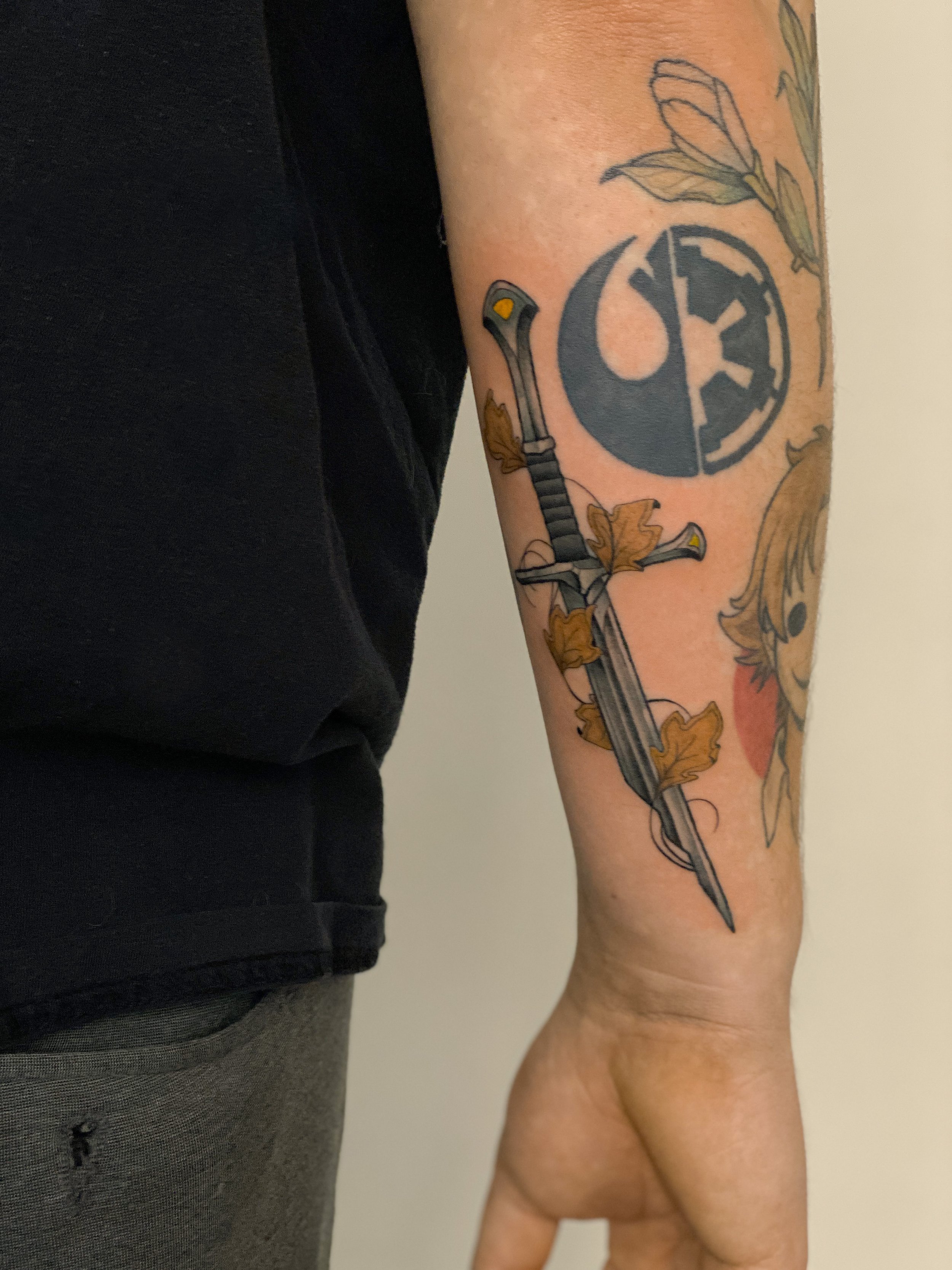 Tatovering af et sværd med forbore, omgivet af brune efterårsblade på underarmen.