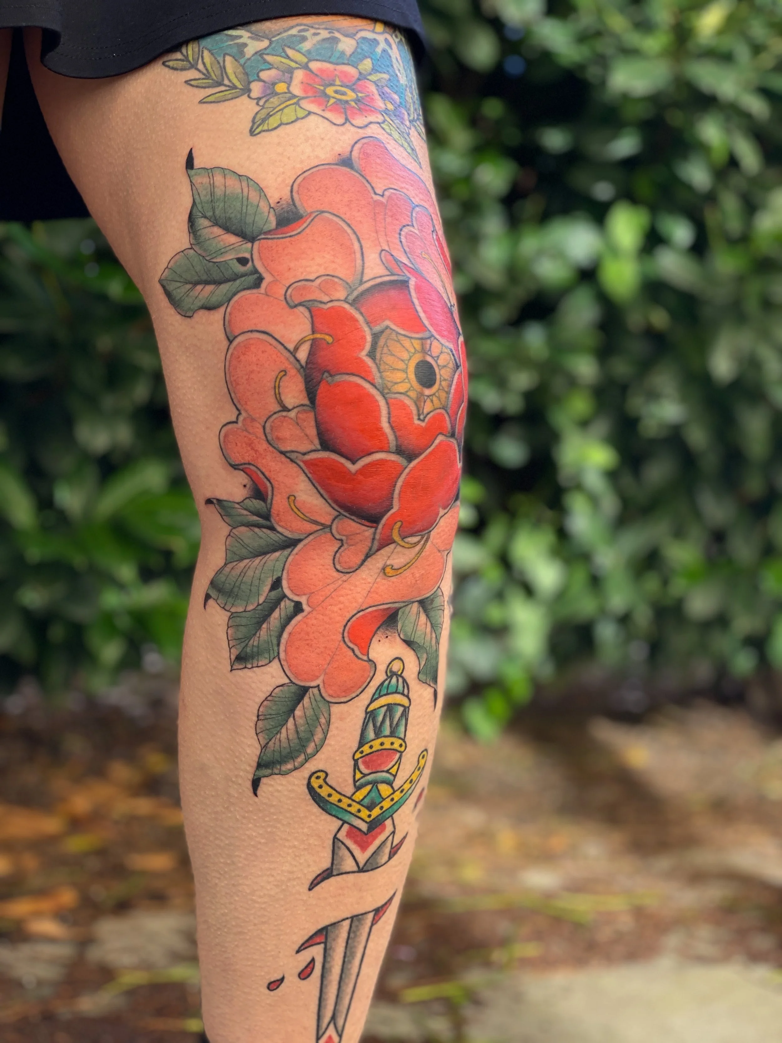 Tattoo på benet med en stor blomsterkirke i rød-orange farve, bladgrønne blade, et sværd med dekorative elementer og nogle små bloddråber.