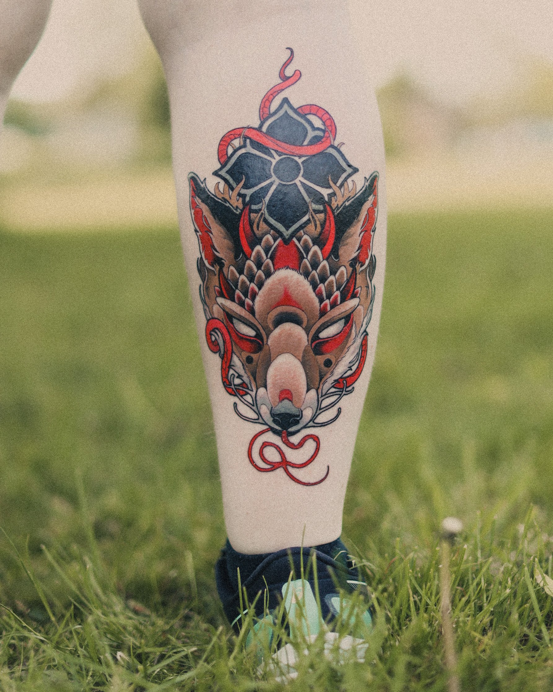 Tattoo af en japansk-inspireret ræv med blomster og snirkler på en persons ben, der står på græs.