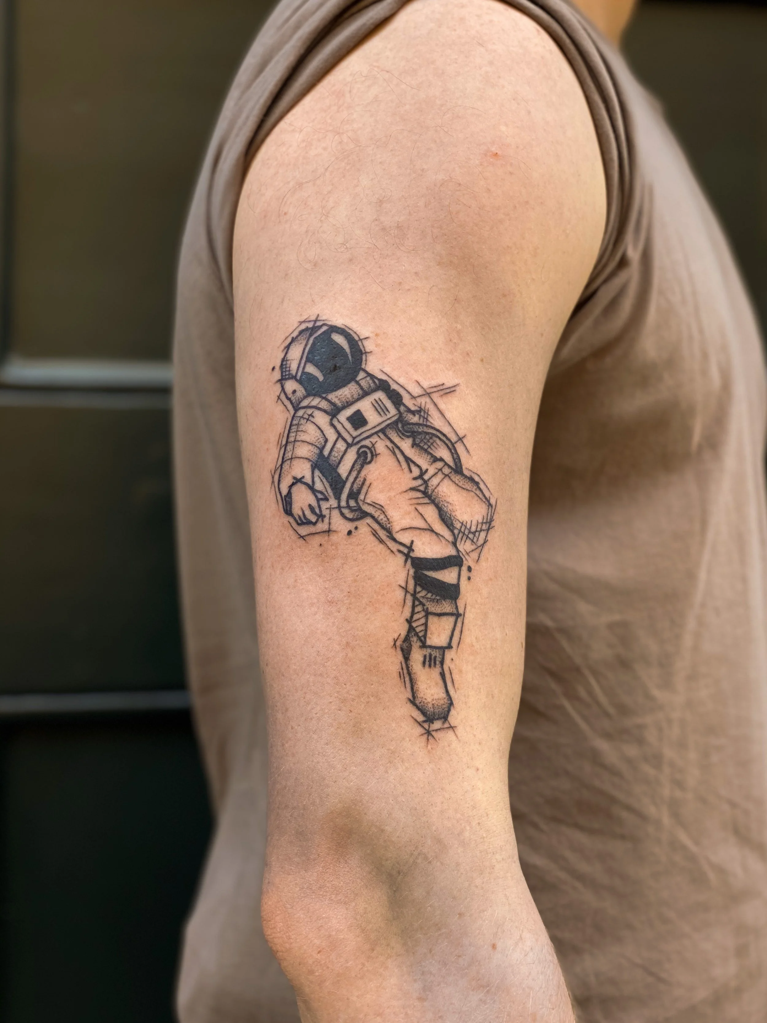 Tegnet tatovering af en astronaut på en persons overarm, designet i en skitse-lignende stil.