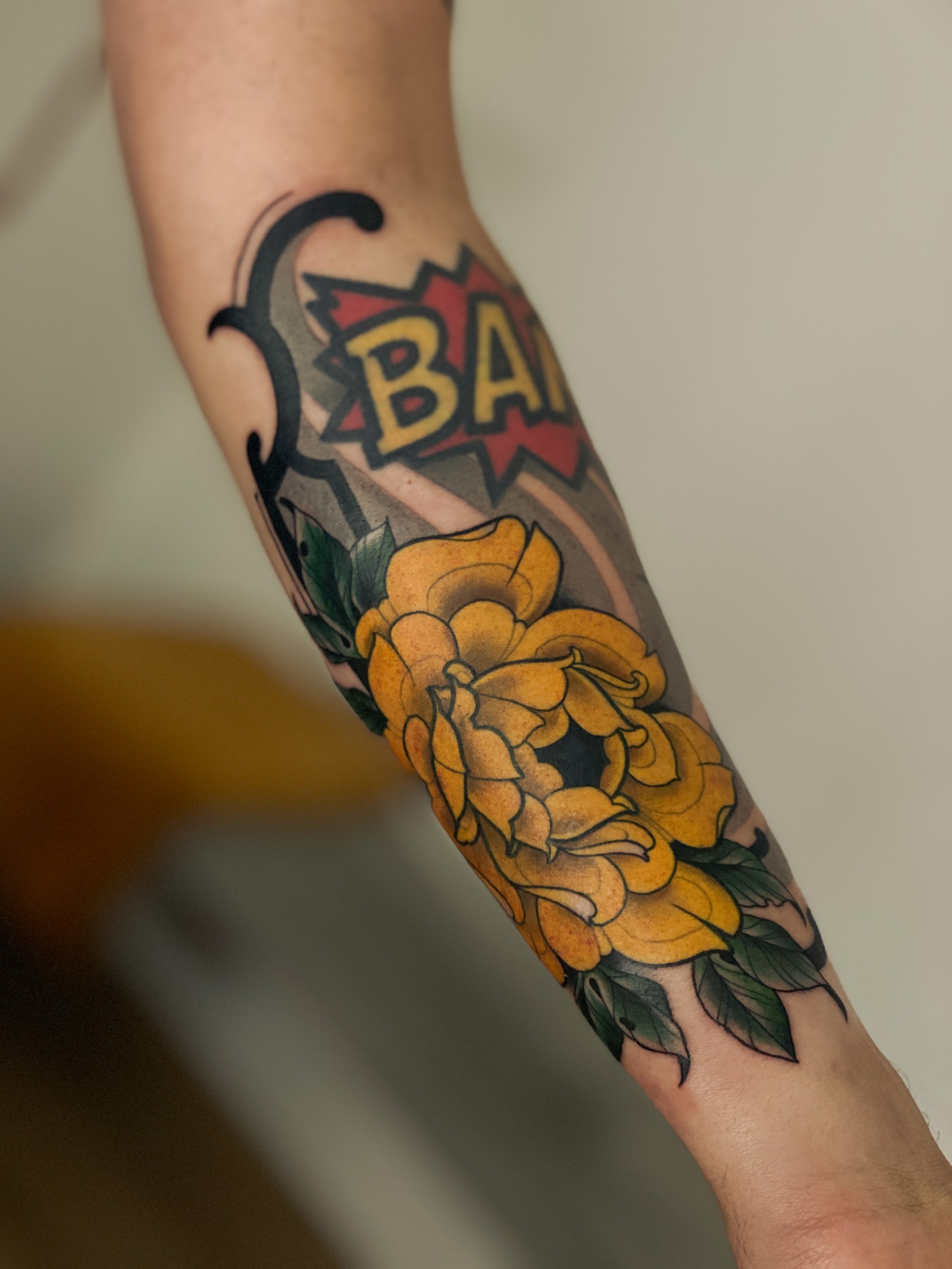 Tatovering af en stor gul blomst med grønne blades og et ord, der ligner 'BANG', i en tegneserie stil.