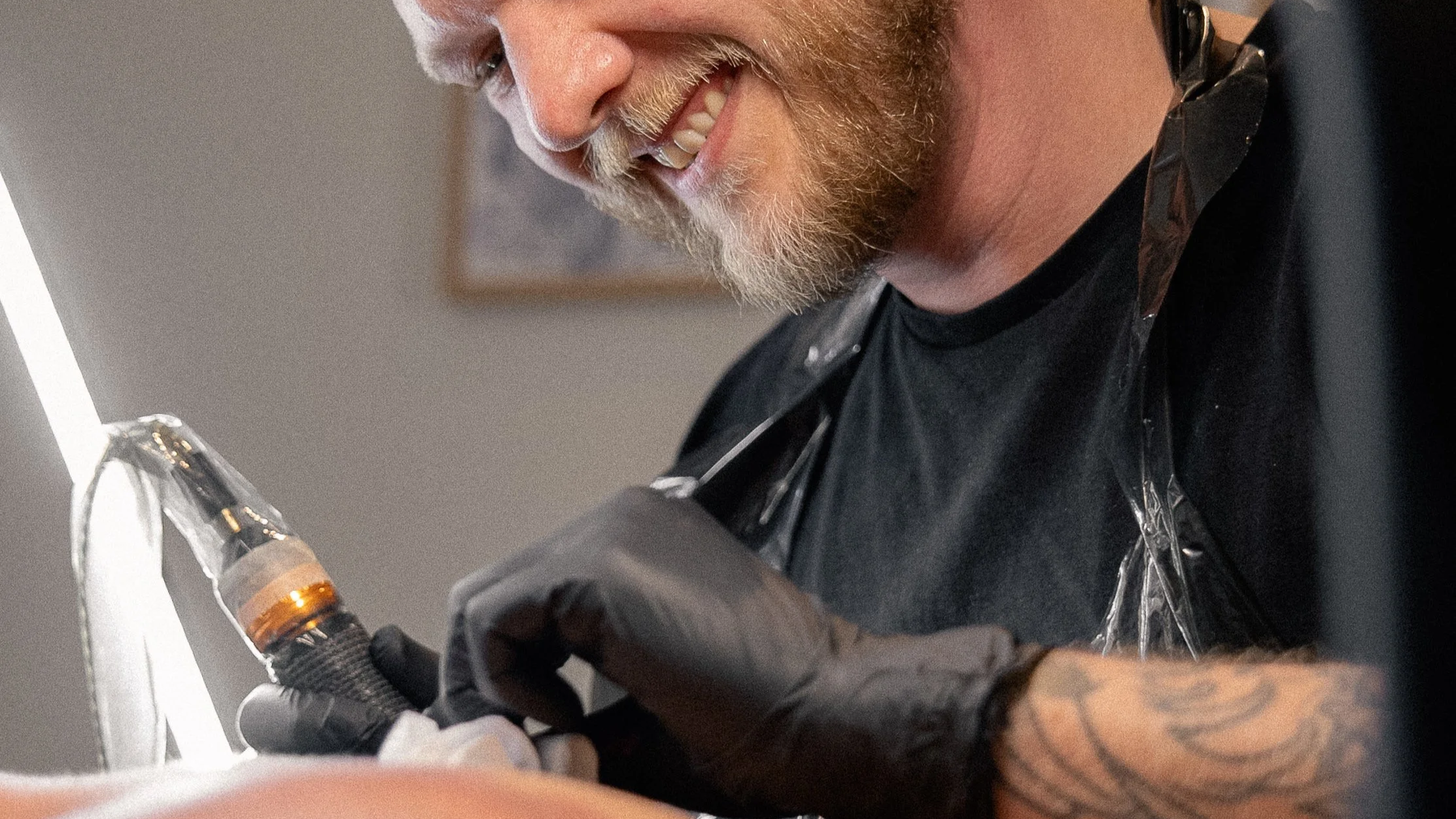 En mand med skæg, der smiler, mens han arbejder med en tatoveringsmaskine. Han bærer sorte handsker og en sort plastikbeskyttelse over skuldrene.