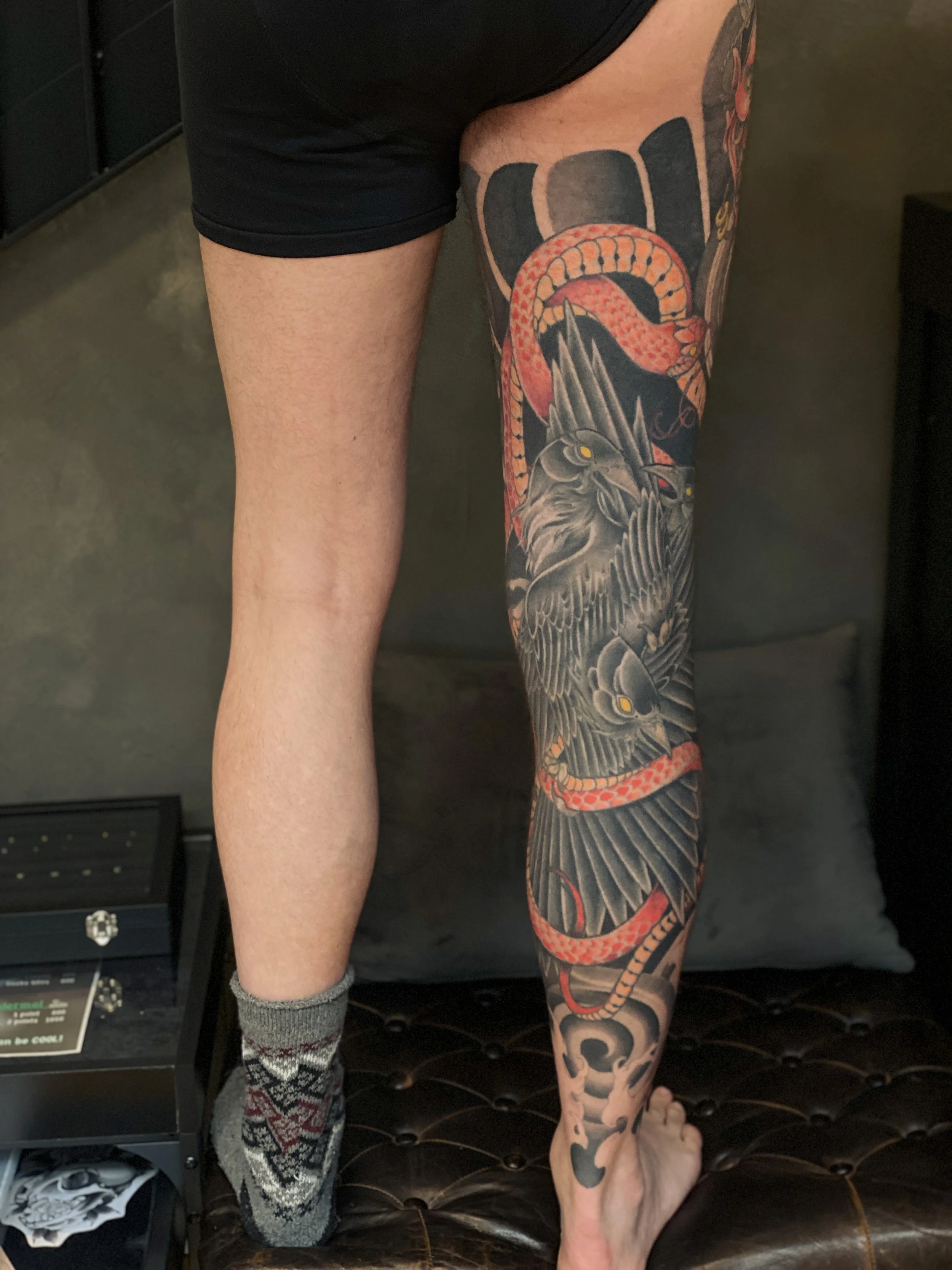 En persons ben med en stor tatovering af en kæmpe ugle og en rød slange, der snoer sig rundt om benet.