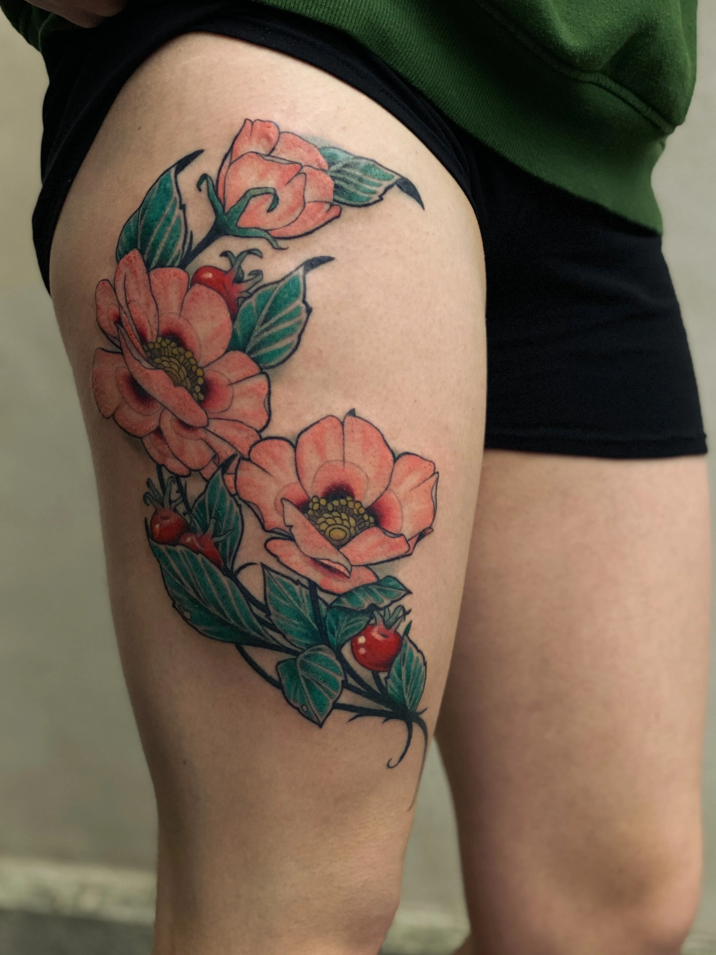 En farvet tatovering af blomster med grønne blade og røde bær på låret.