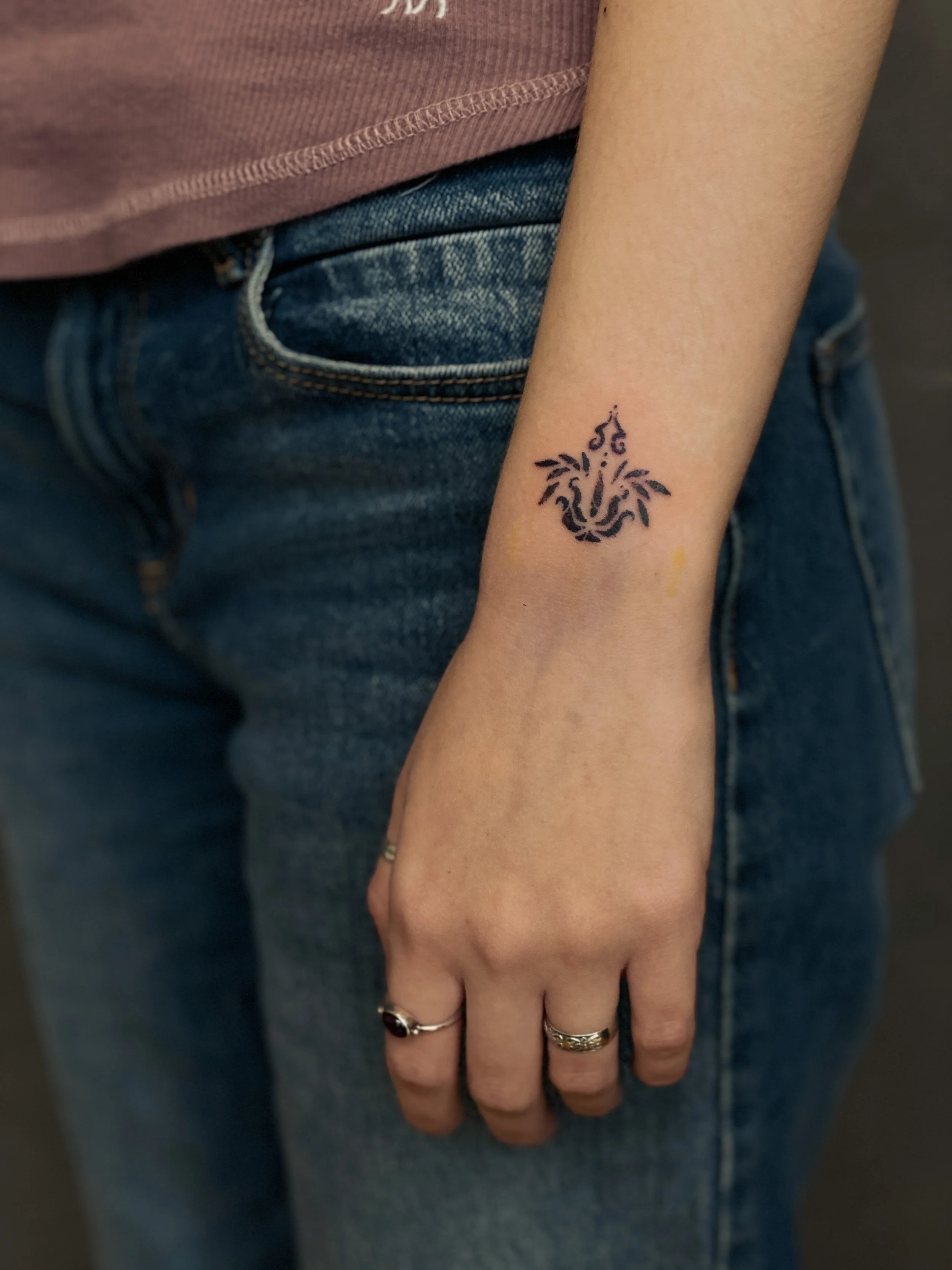 Tatovering af en løve med sort blæk på personens indre håndled, personen har flere sølvringe på fingrene, og de er iført blå jeans.