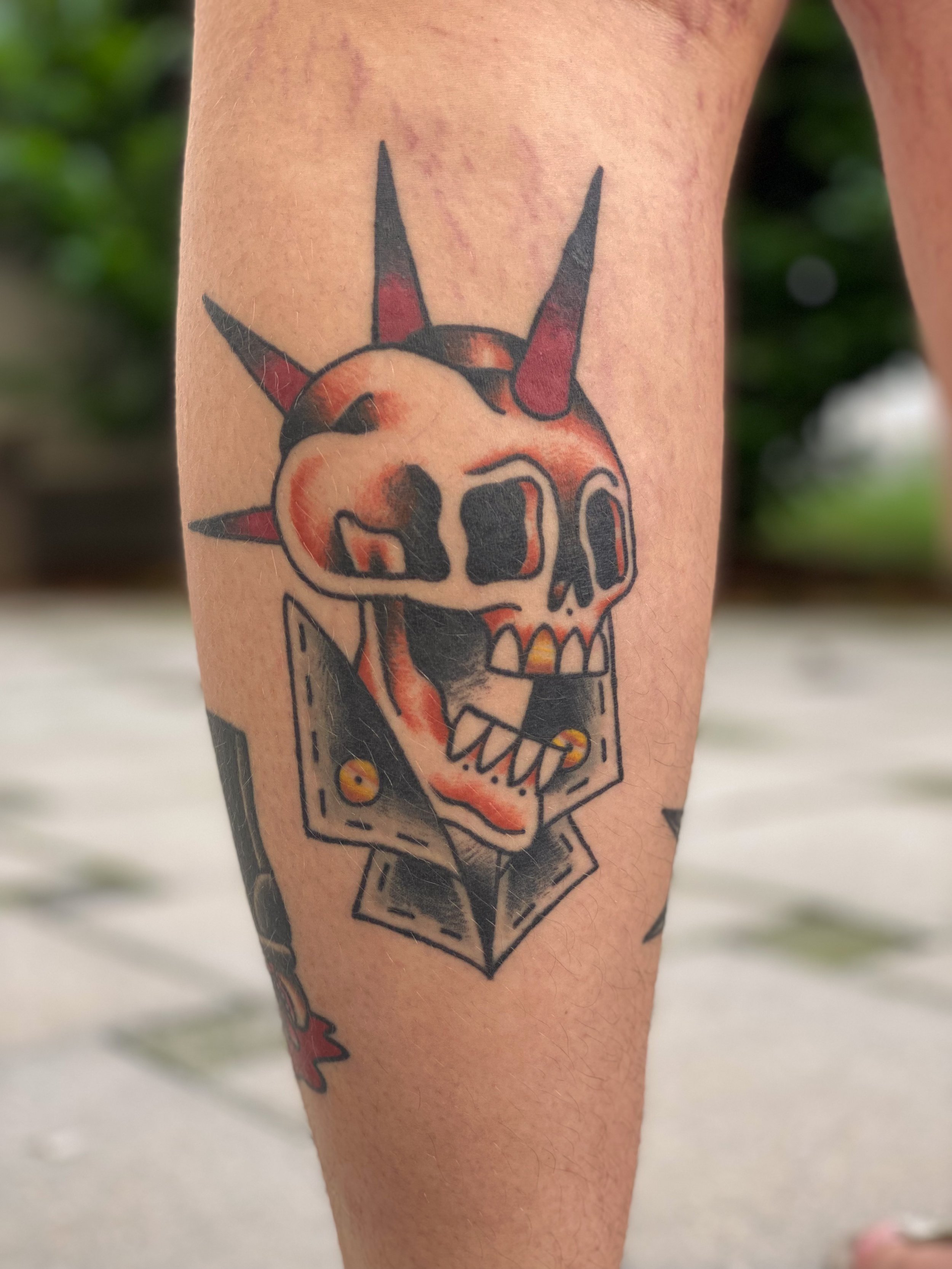 Tatovering af en femkantet skalle med horn, der har en bulldog-udseende, med snit, sort og orange farve, i en sort ramme.