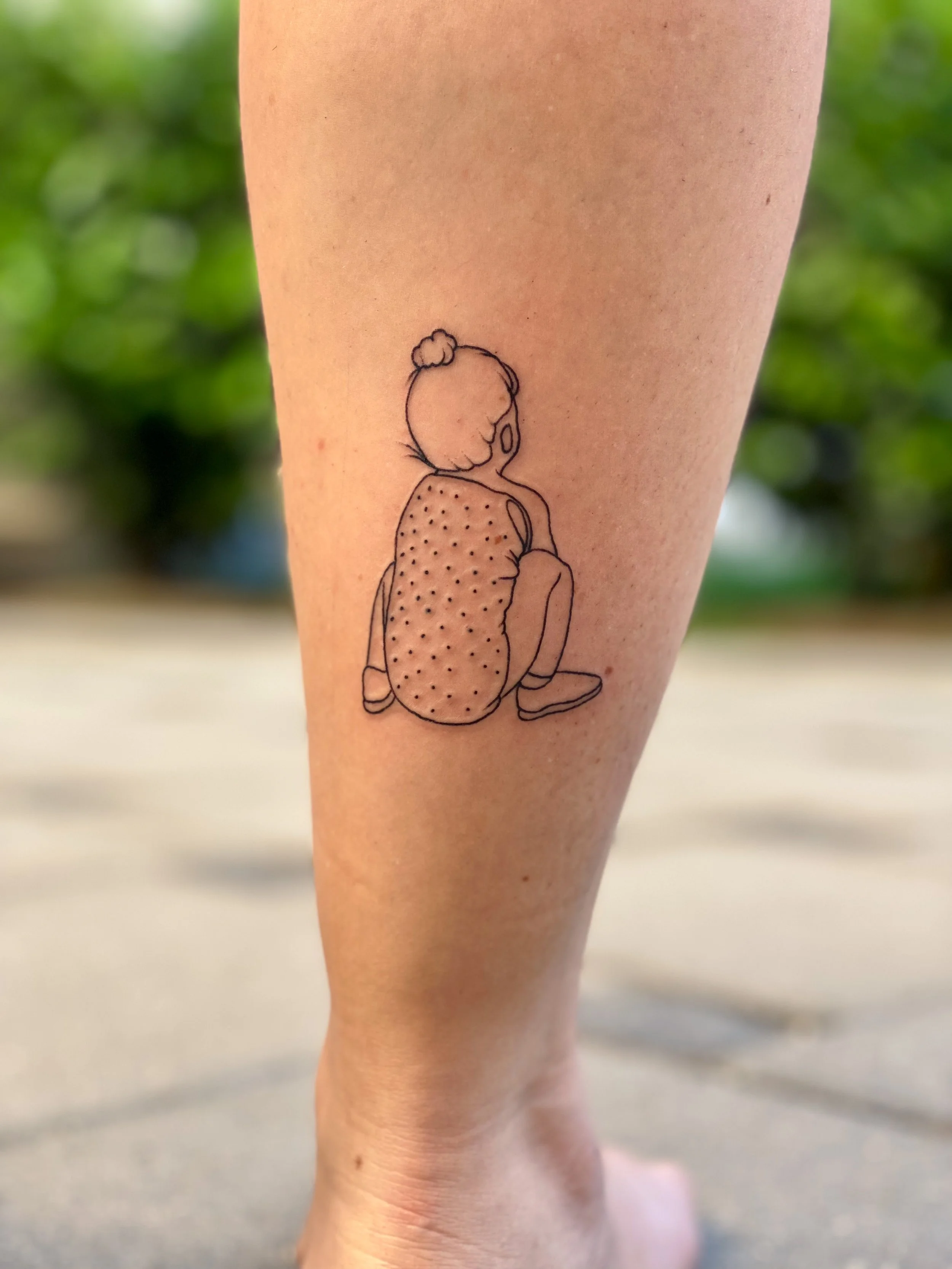 Tatovering af en enkel stregtegning af en baby, der sidder med ryggen til, set fra bagfra, med hår i en lille knold og en klud over skulderen.