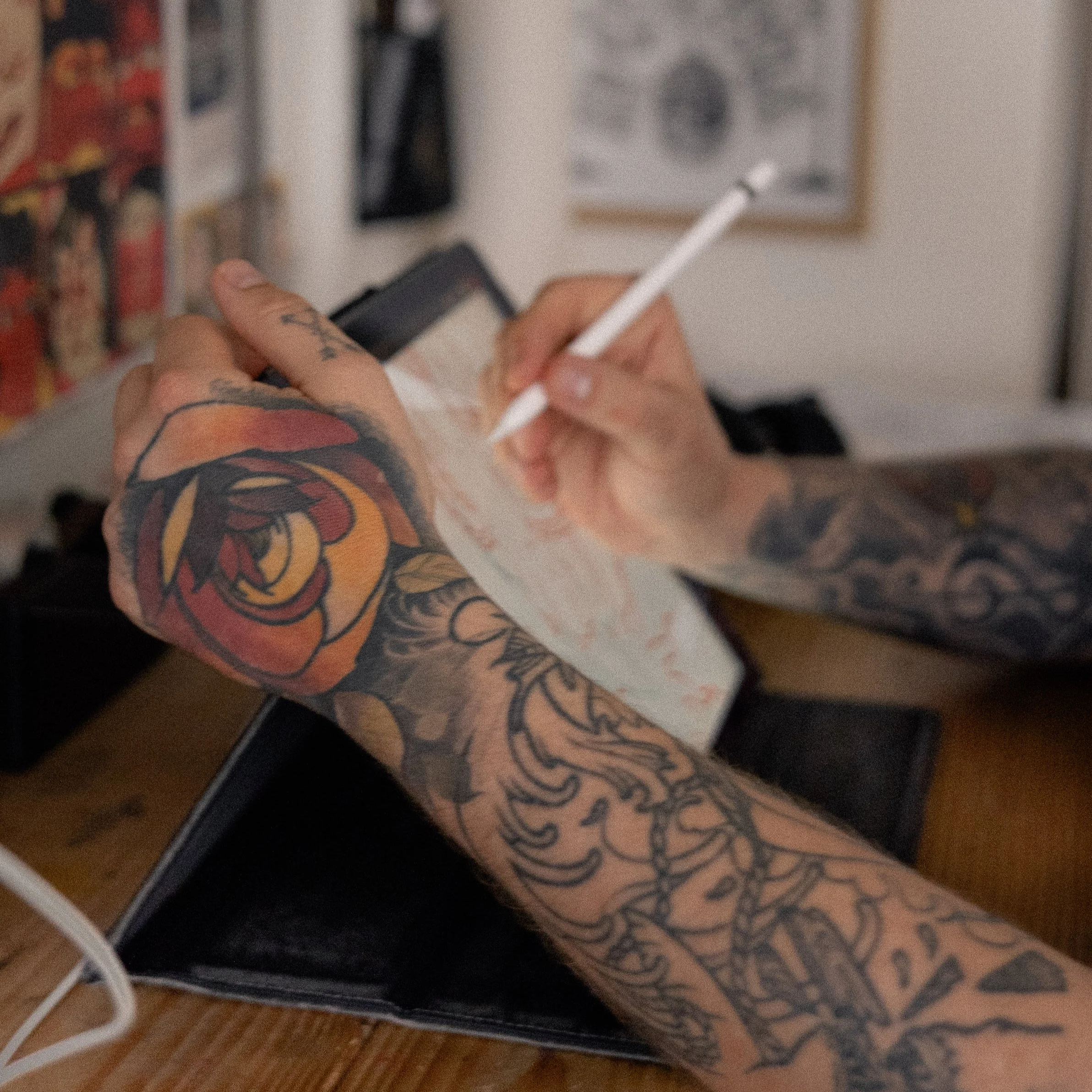 Tatoverede hænder skriver med en stylus på en digital enhed, omgivet af tatoveringer.