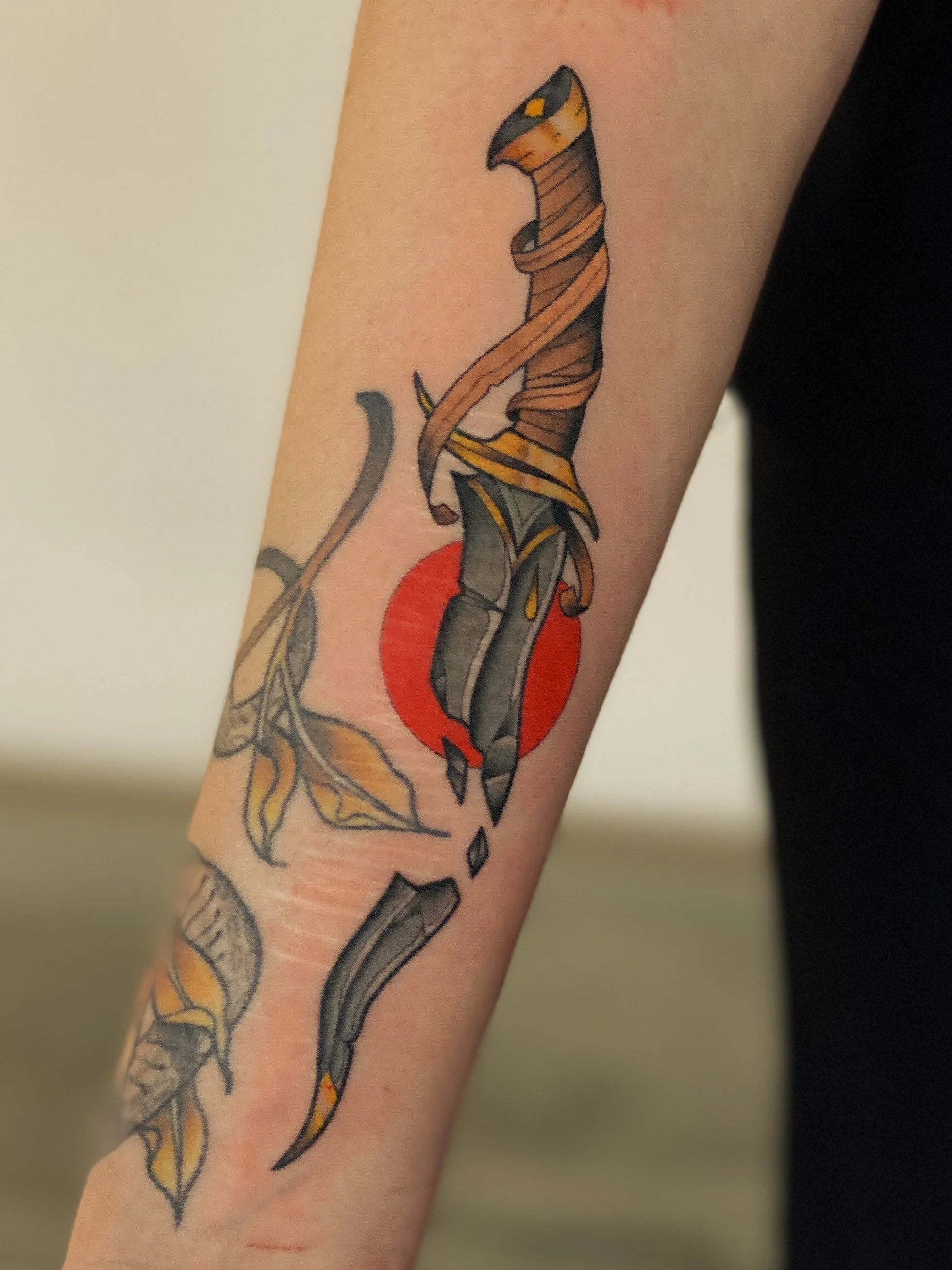 Tattoo af en sværd med en klinge, omgivet af blade og blade, foran en rød cirkel på underarmen.