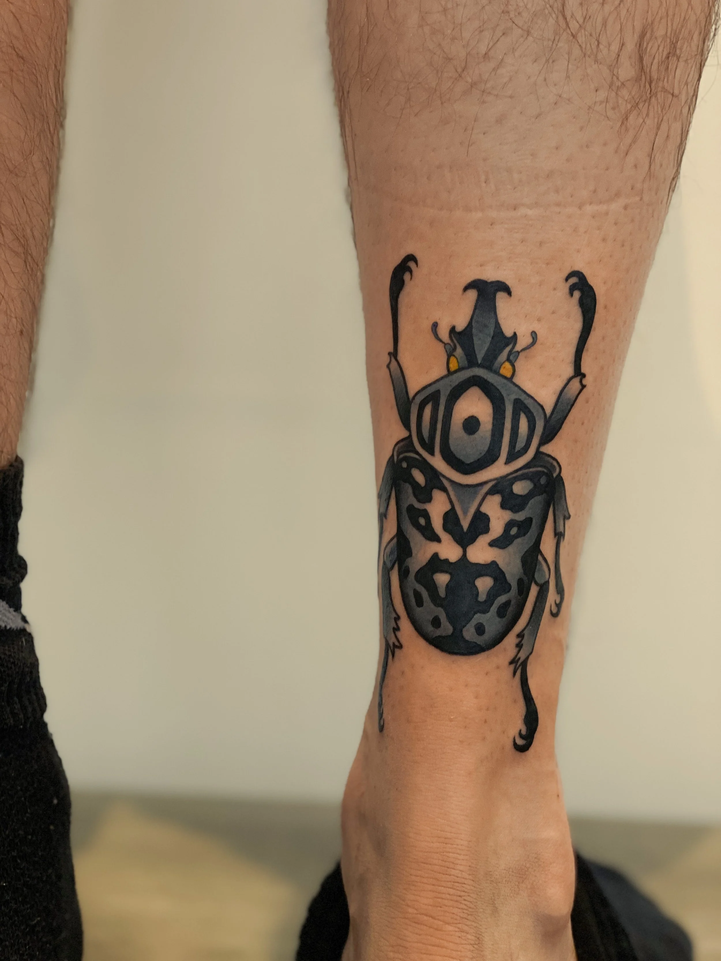 Tattoo af en styliseret bille af et insekt med sorte og grå nuancer og gule øjne på persons ben.