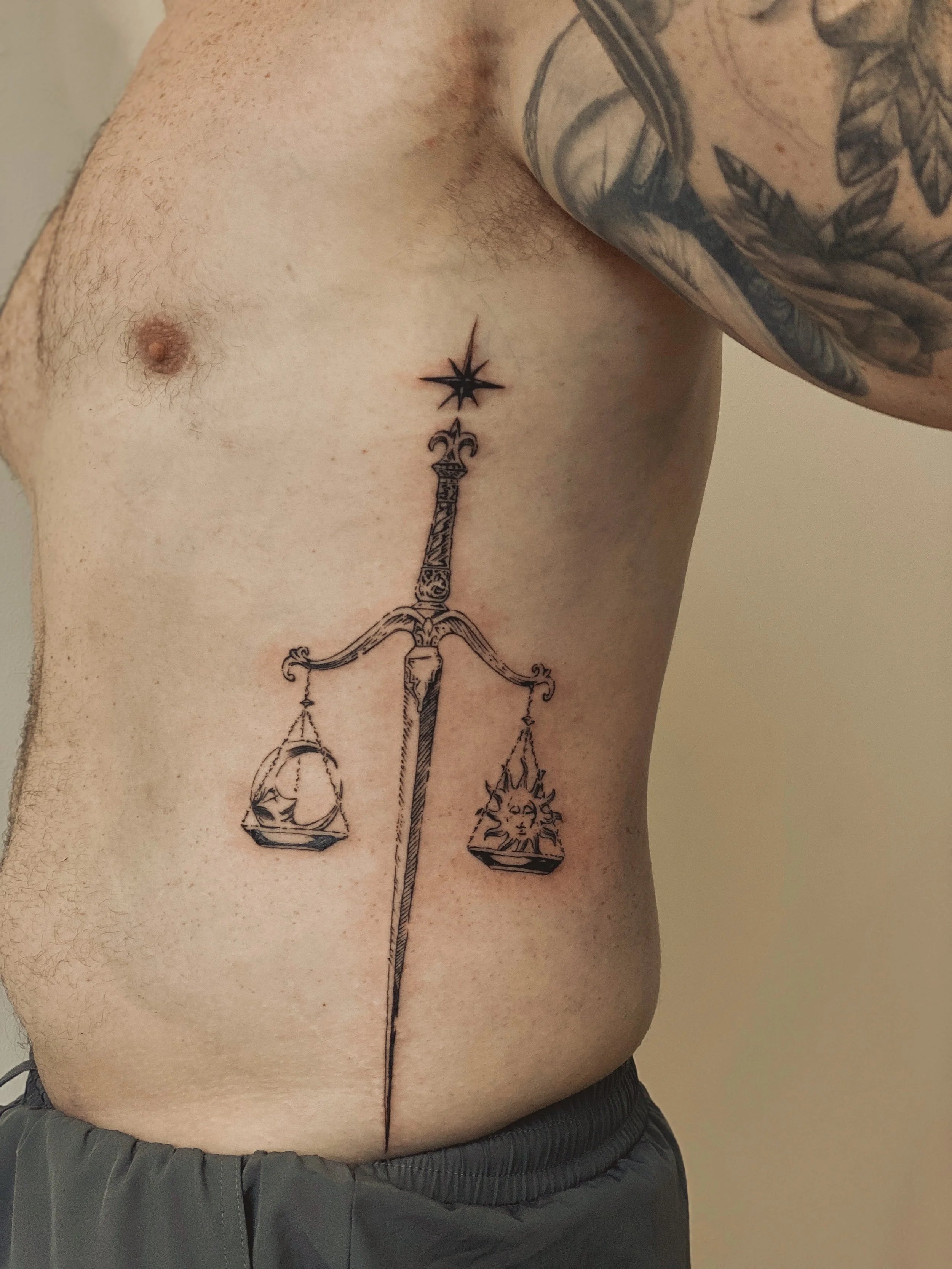 Tatovering af en vægt med en sværd som stang, med en sol med ansigt på den ene vægtskål og en måne med ansigt på den anden.