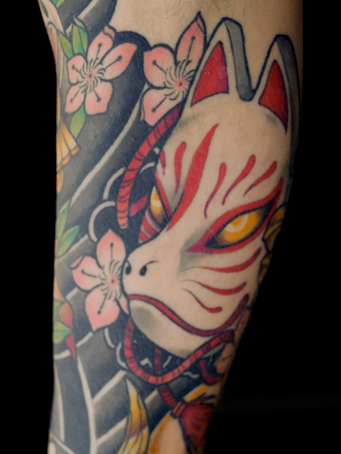 Tatovering af en japansk inspireret kat med gule øjne og røde striber, omgivet af lyserøde kirsebærblomster.