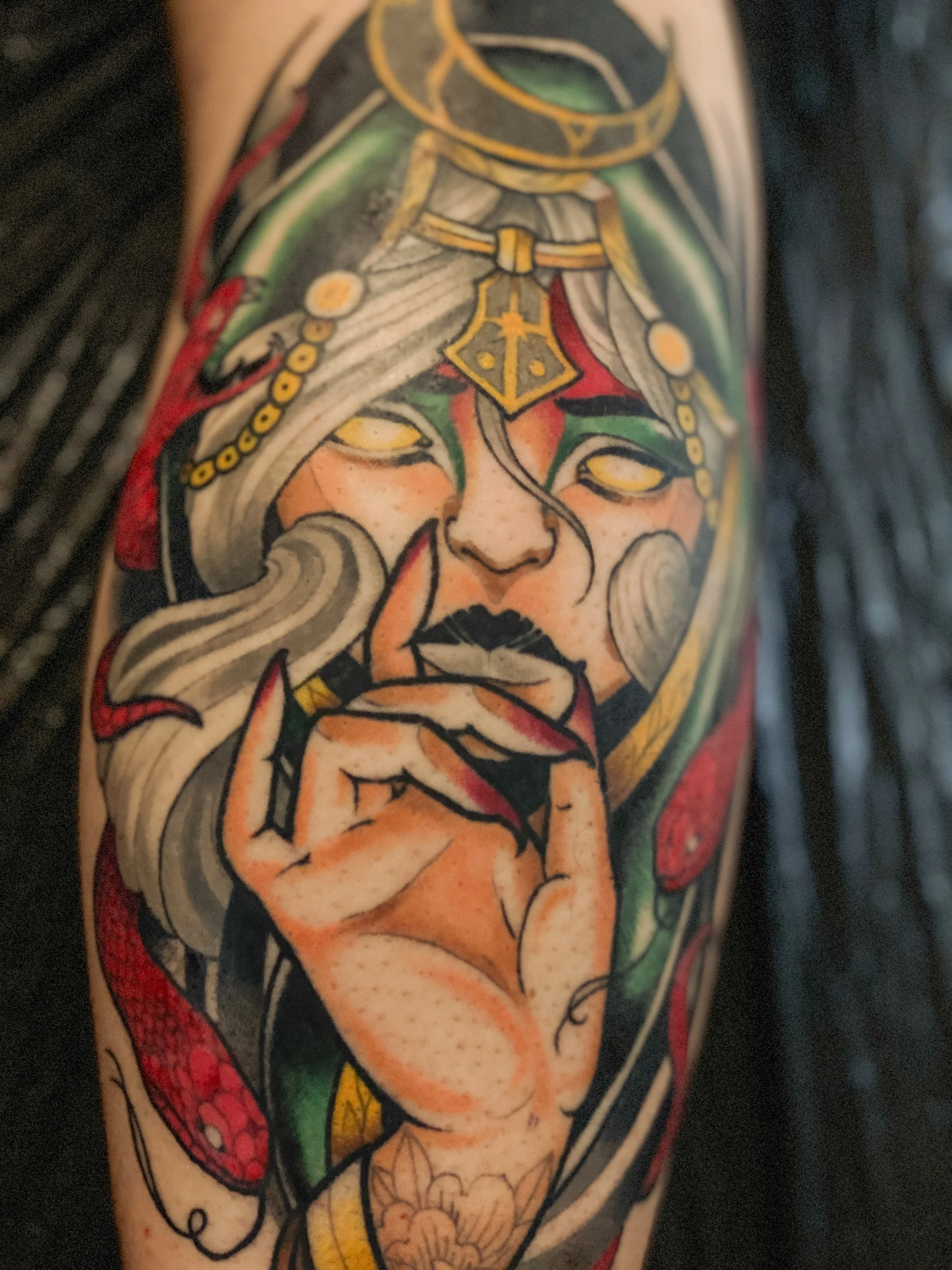 Tatovering af en kvindelig figur med hand i ansigtet, med grøn, rød og sort farve, detaljeret med smykker og mønstre.