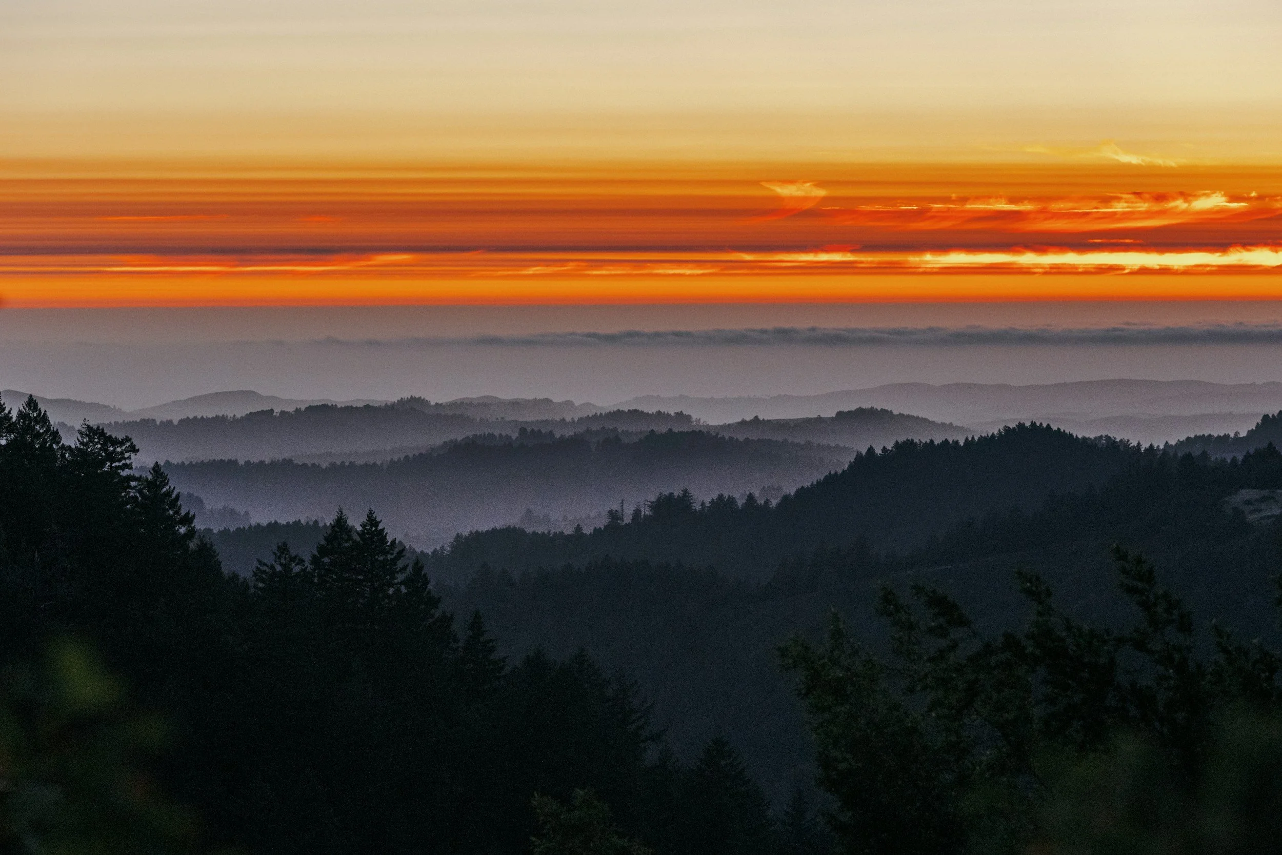 A sunset over a mountain range with layers of misty trees and clouds in the sky.