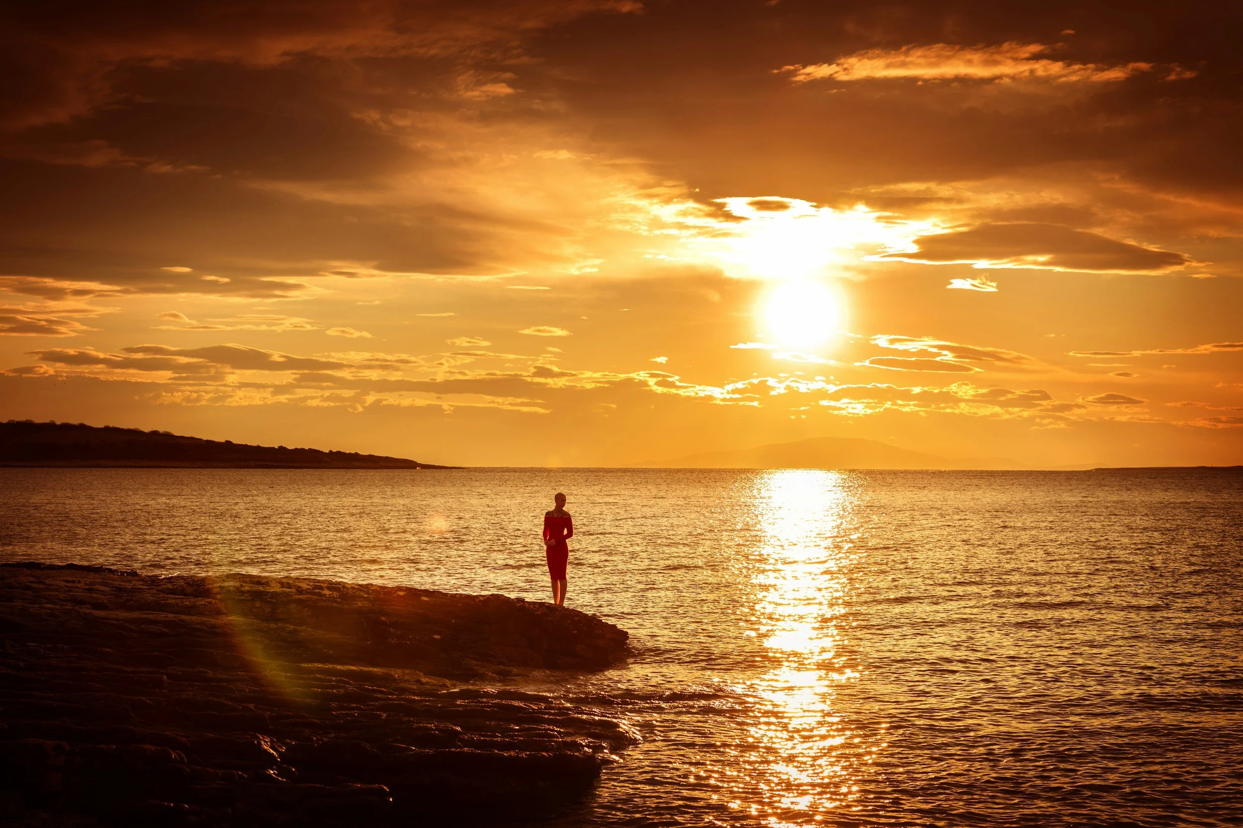 A person standing on a rocky shore during sunset, with the sun low in the sky and clouds scattered above, reflecting on the water.