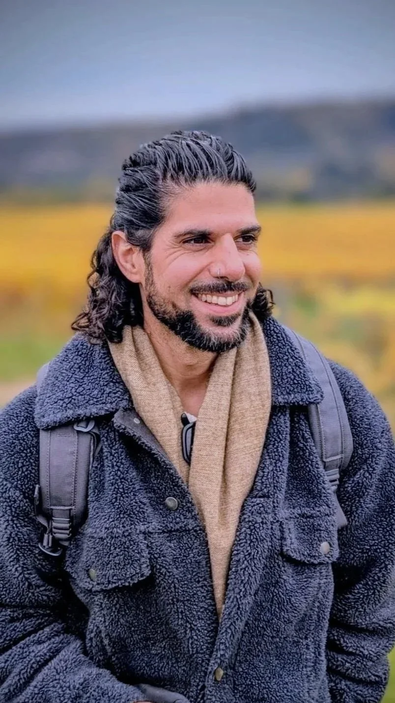 A smiling man with long dark curly hair and a beard, dressed in a fleece jacket and a tan scarf, standing outdoors with a blurred background of yellow and green fields and mountains.