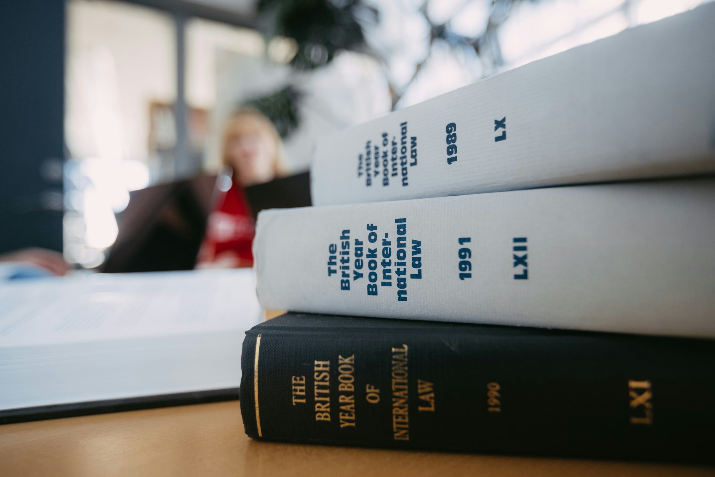 Close-up of legal books on international law, with titles indicating years 1989 and 1991, and a woman with blonde hair and red shirt in the blurred background.