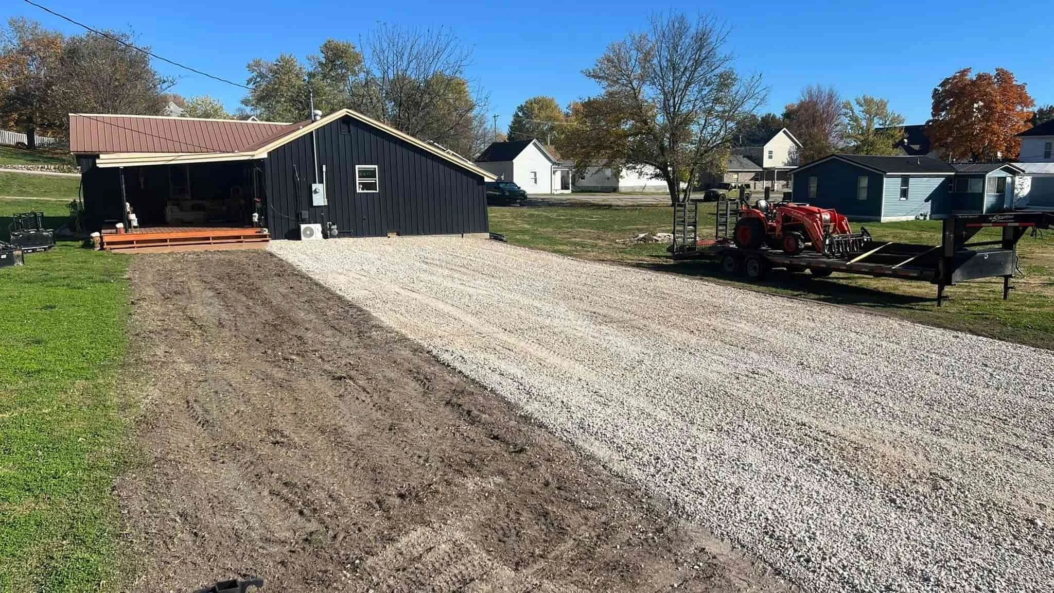 A newly gravelled driveway leading to a dark blue shed with a metal roof, situated in a backyard with grass and trees, under a clear blue sky.