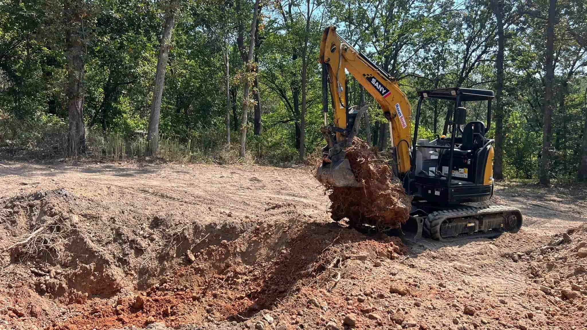 A small yellow excavator is digging into the dirt in a forested area, lifting a large clump of soil and roots.