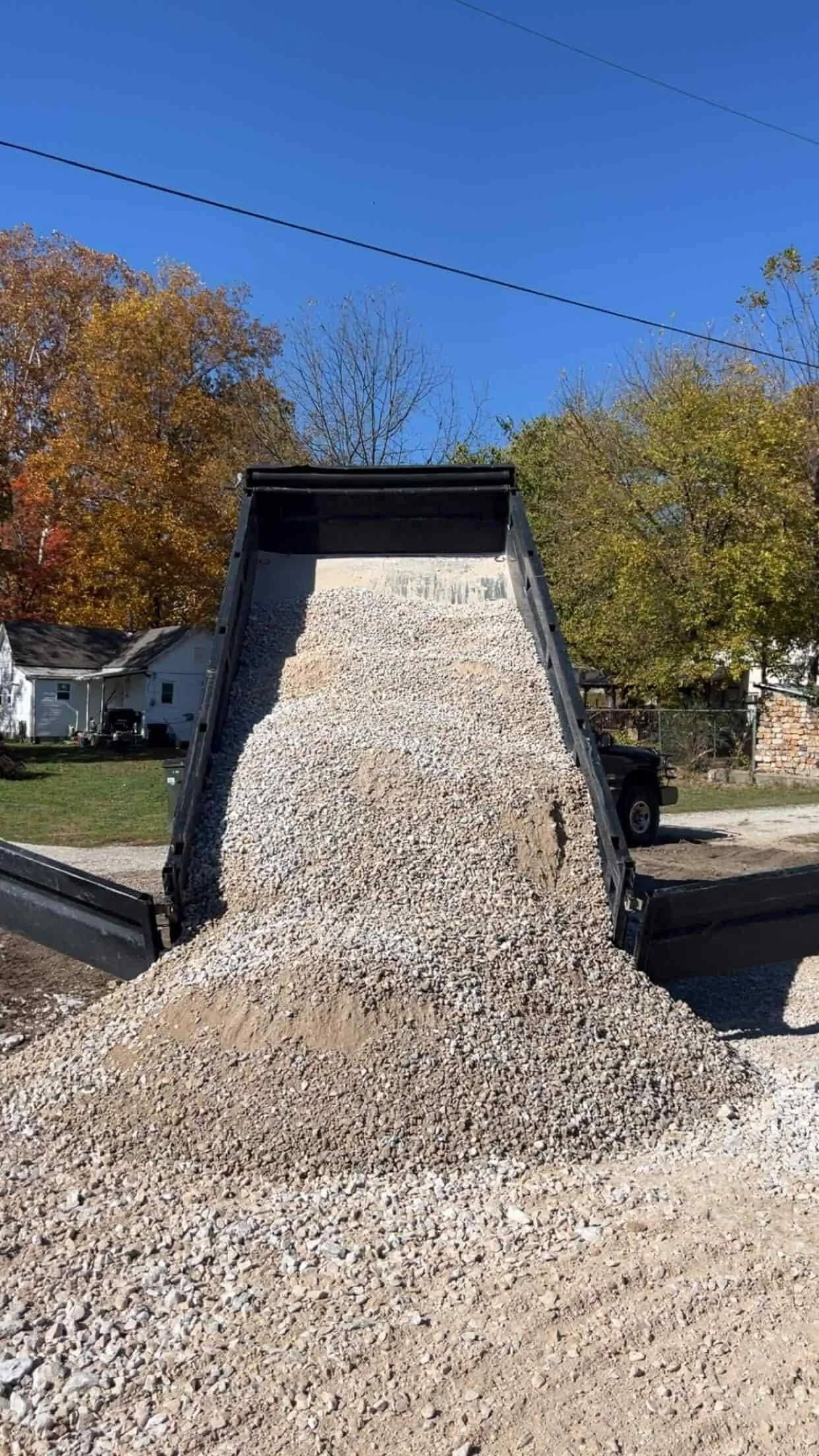 A dump truck unloading gravel onto the ground in a residential neighborhood with trees and houses in the background under a clear blue sky.