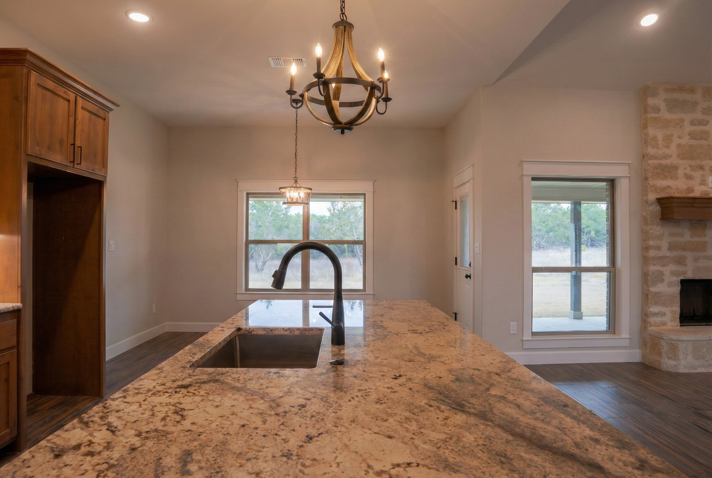 Professional real estate photography of a luxury kitchen featuring a large island, built-in refrigerator recess, and upscale pendant lighting. Long-view perspective includes the sunlit breakfast nook and a glimpse of the living room fireplace. Servin