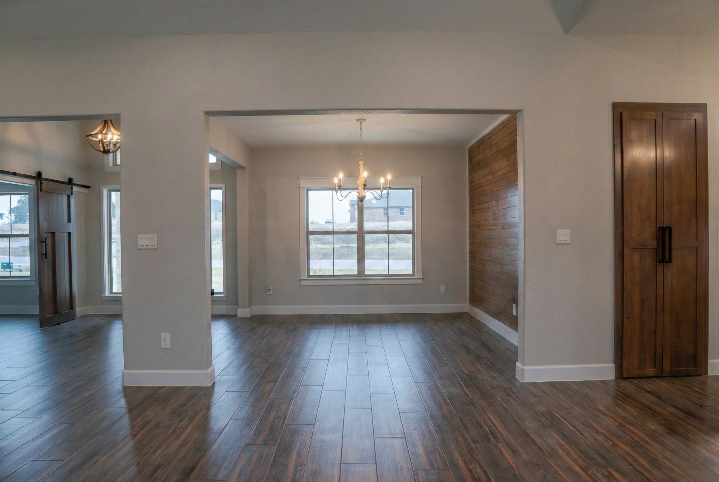 Professional real estate photography of a formal dining area separated by architectural pillars, featuring a wooden accent wall and upscale pendant lighting. View includes a glimpse of the home office barn doors and beautiful wooden pantry doors. Ser