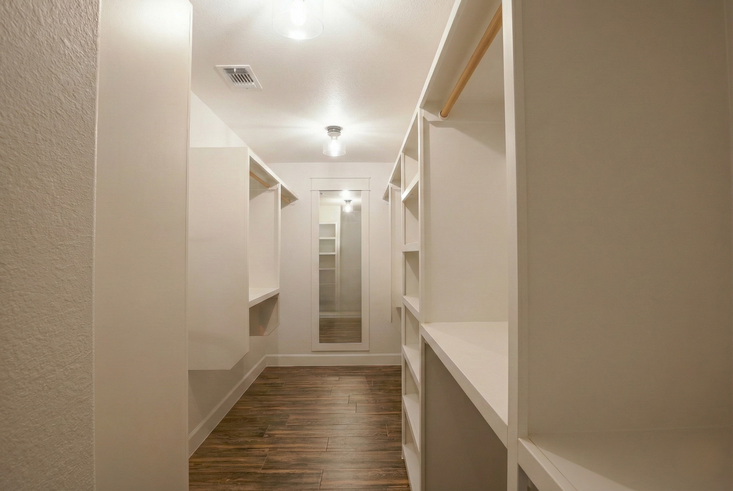 Professional real estate photography of a spacious luxury walk-in closet in a vacant 3,500 sq ft home. Long-view perspective featuring custom built-ins on both sides, ample storage, and a full-length mirror on the far wall. Serving Austin, Round Rock