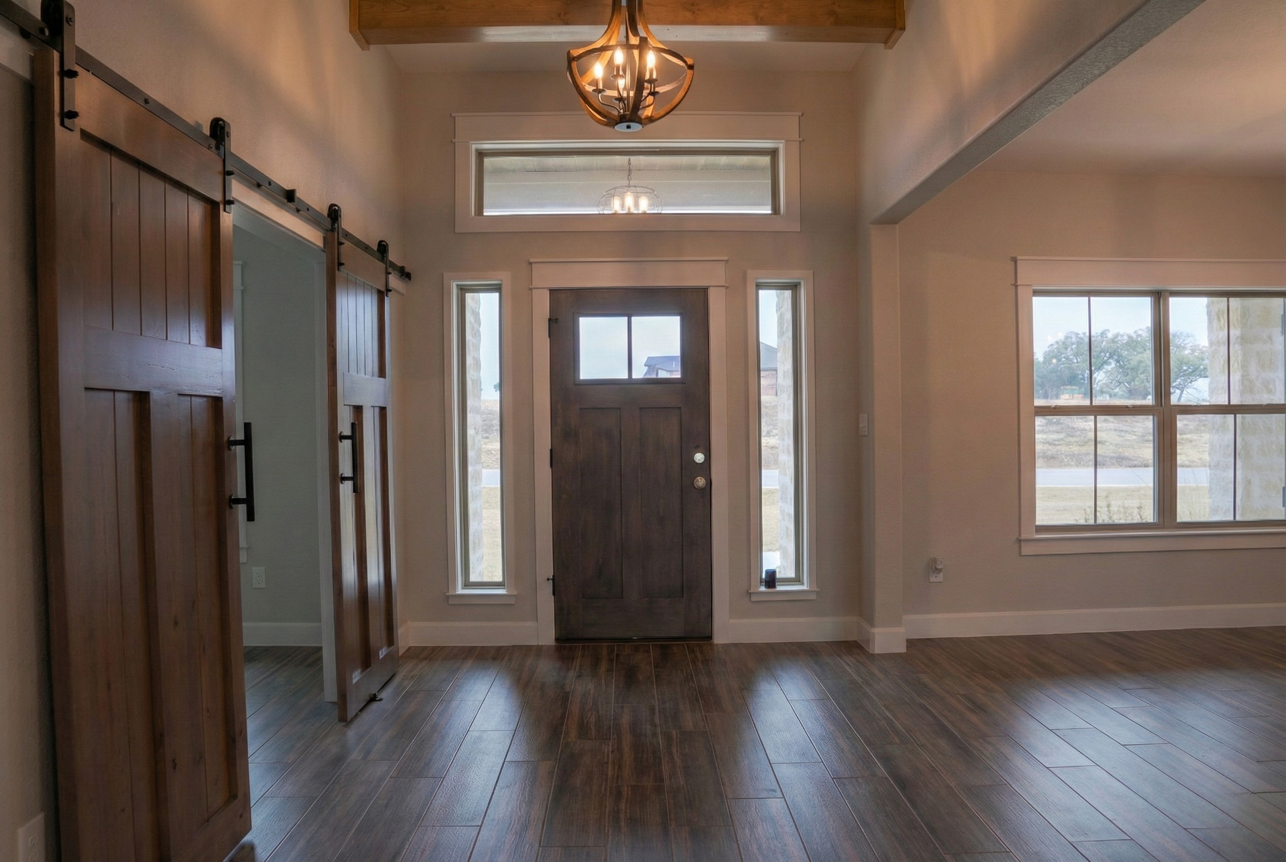Professional real estate photography of a luxury home entryway featuring high ceilings, a beautiful chandelier, and a barn door style entrance to the home office. View includes a glimpse of the dining room with ample windows and natural light. Servin
