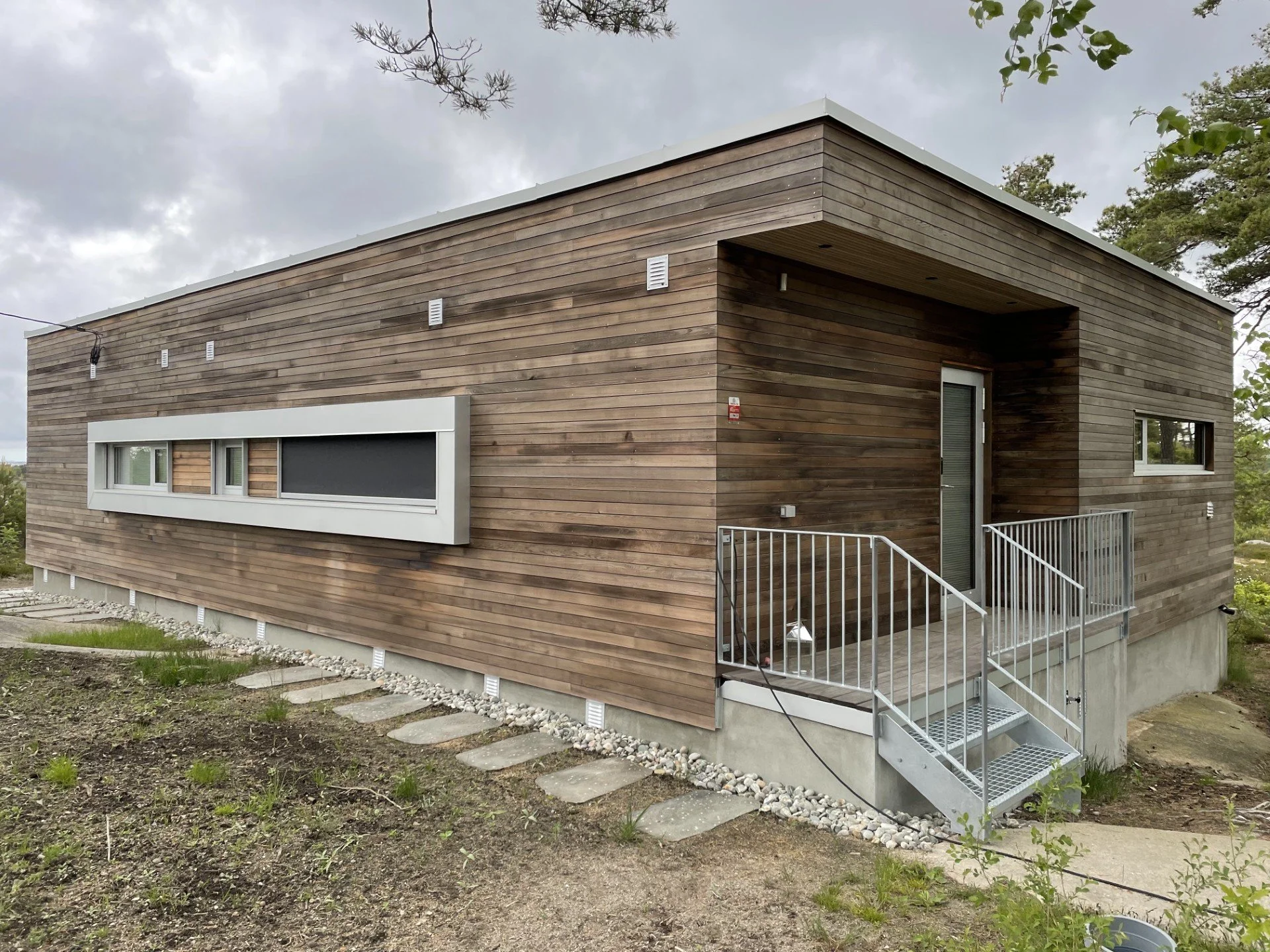 Moderne trehus med flat tak, hvite rammer rundt vinduer, inngang med metalltrapp, omgitt av utendørsområde med stein og gress, under en overskyet himmel.