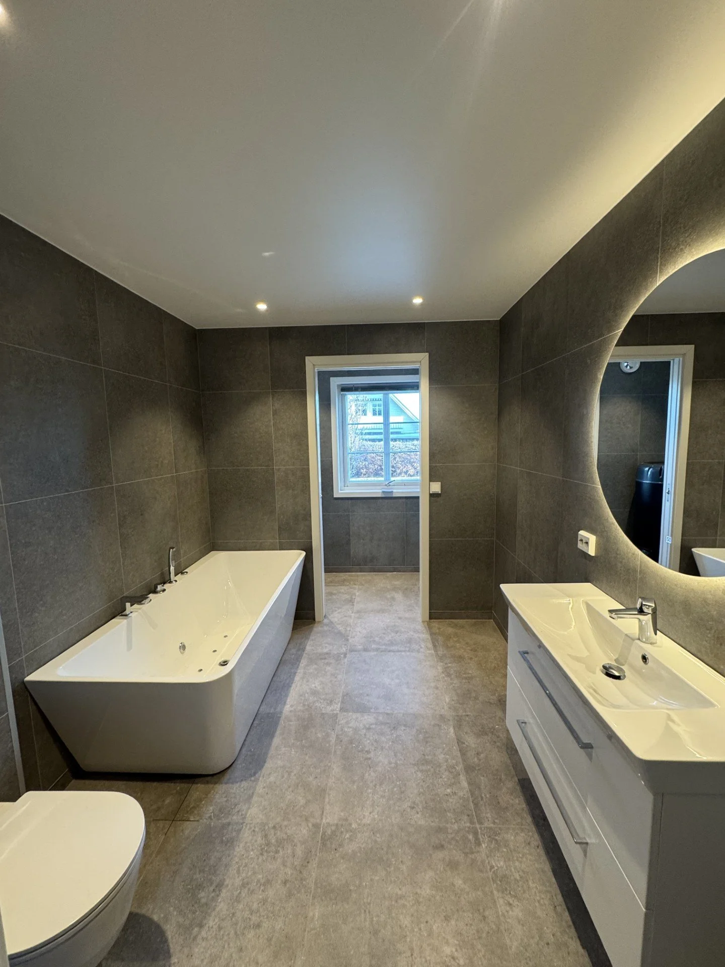 Moderne bad med hvit badekar, stor speil over vask, grå vegg- og gulvfliser, vindu, hvit vask med skuffer.