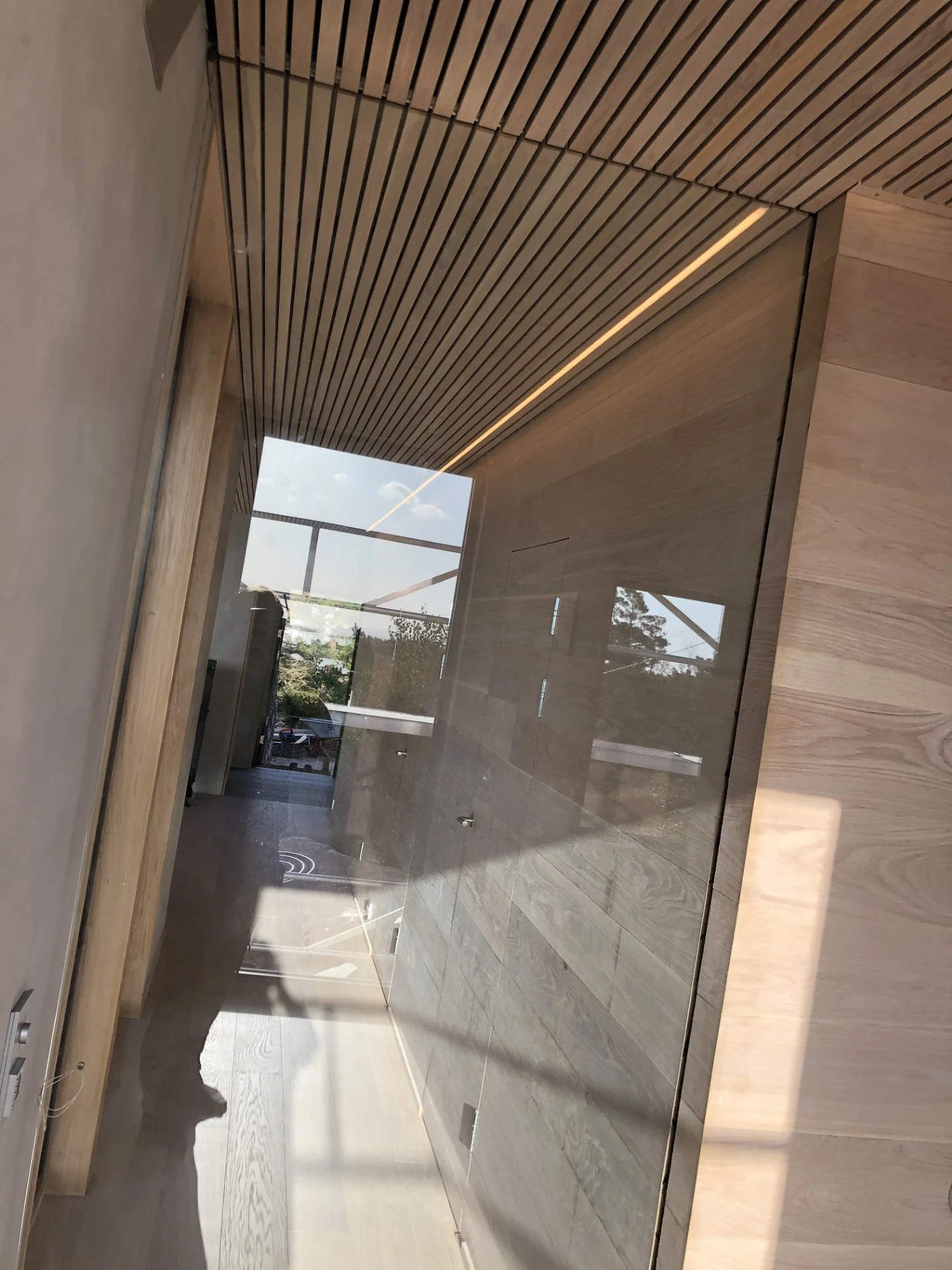 Innvendig rom med treribbet tak, lyse trebordgulv og en stor glassvegg mot utsiden. Refleksjoner i glasset viser det utendørs miljøet.