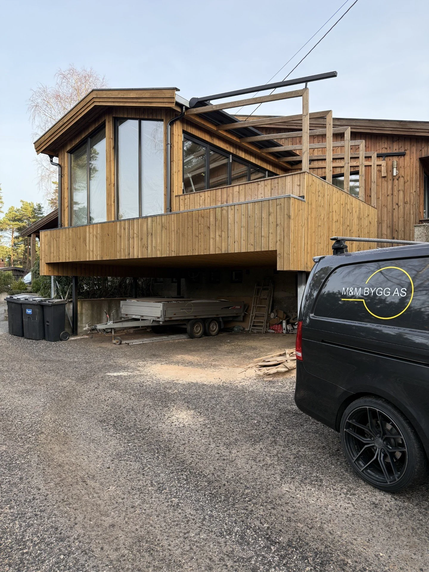 Nylig byggebolig med tredekk og store vinduer, en svart firmabil med logoen M&M BYGG AS er parkert utenfor, og det er byggeutstyr og søppel på tomten.