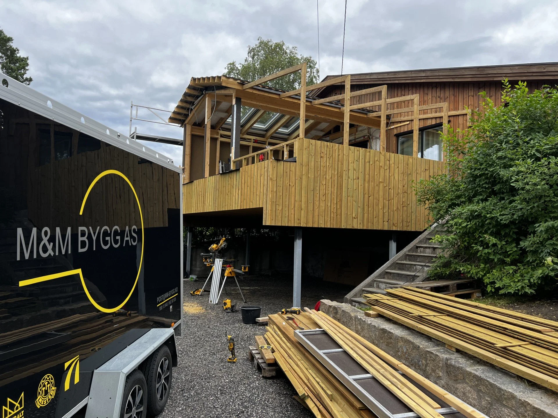 På en byggeplass holder de på å bygge en terrasse på en moderne bolig med treverk. Det er byggemateriell, verktøy og byggutstyr rundt området.