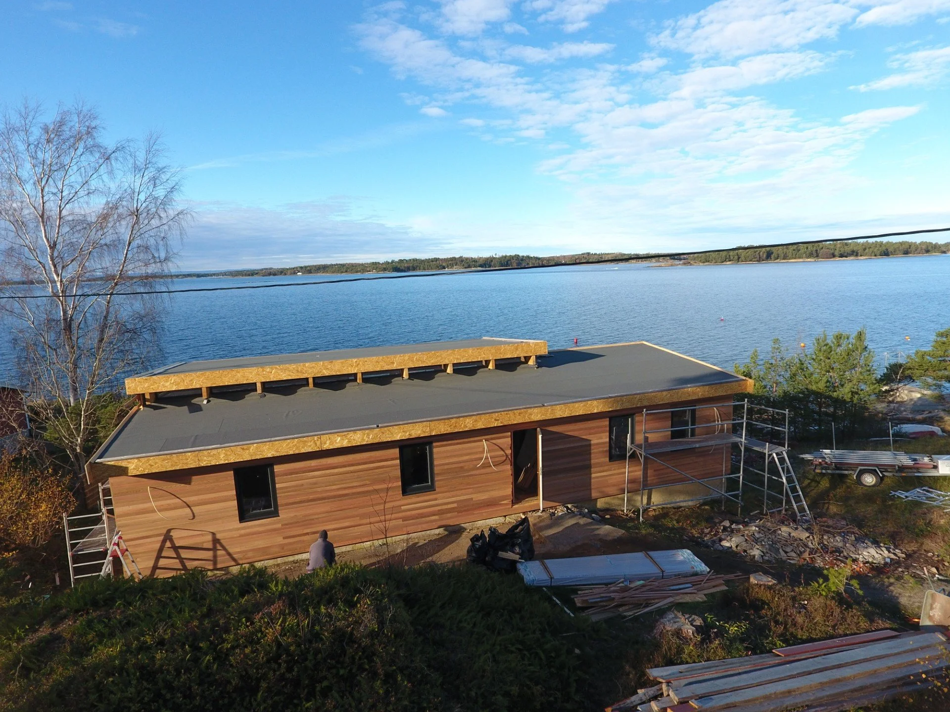 Et hus under bygging ved en vannkyst, med en sandstrand og trær i bakgrunnen, klare blå himmel og sky.