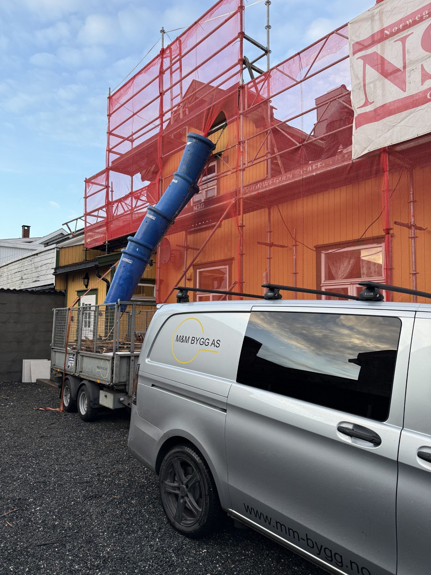 Konstruksjon av et hus med skum og maling, omgitt av midlertidig beskyttelse med skum og en blå transporttube, med en firmabil med logoen "M&M Bygg AS" og nettsiden "www.mm-bygg.no" parkert foran.