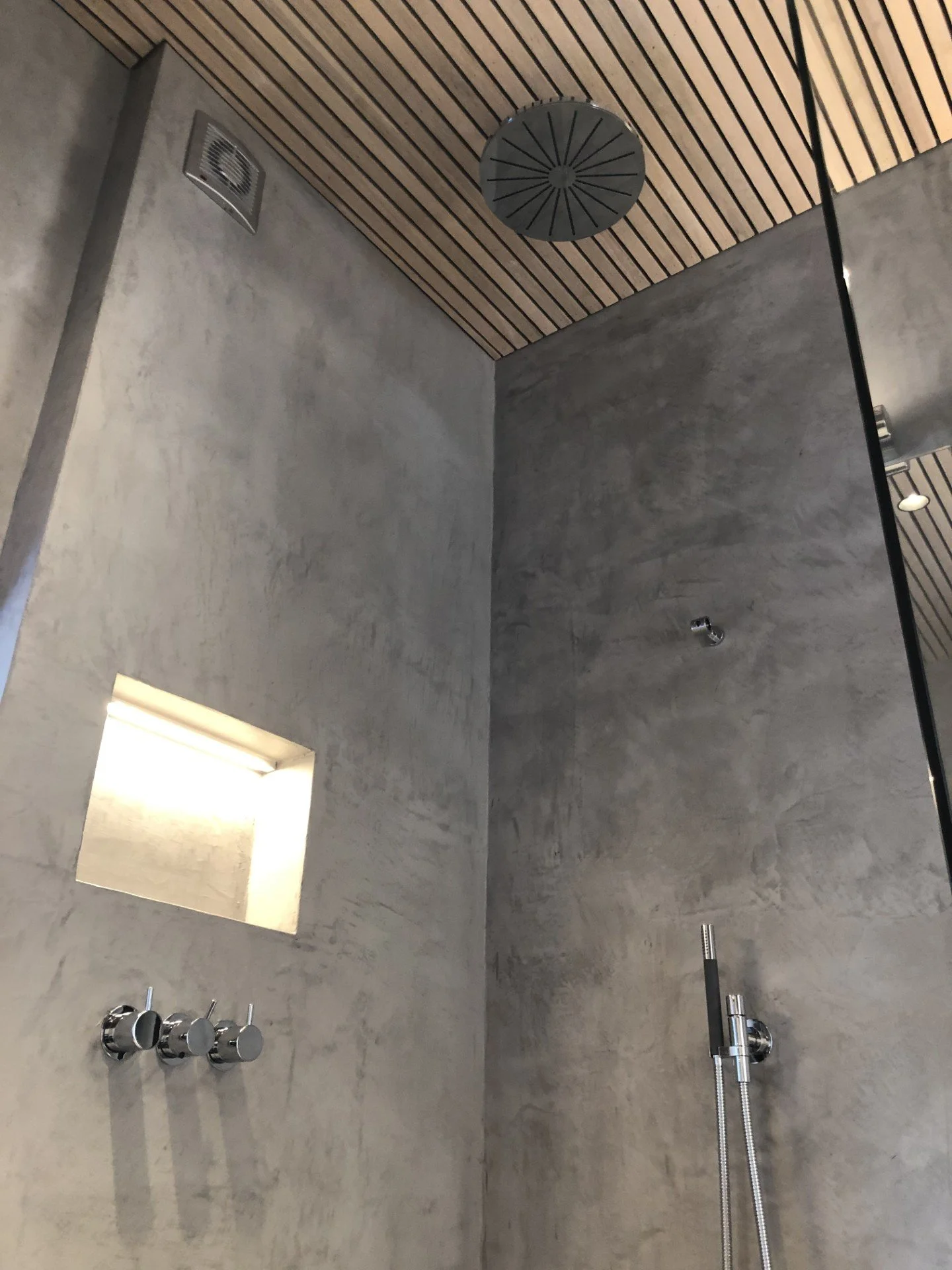 Dusj med grå vegg og trepanel i taket, et innfelt lys over en nisje, en takhånddusj og en vegghengt håndhånddusj.