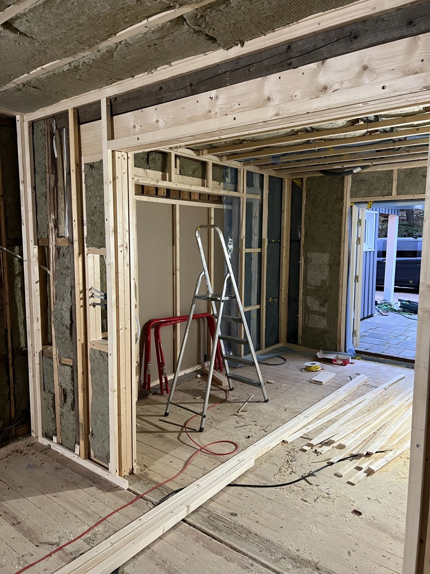 Innvendig byggeplass med treverk og verktøy, klar for å bygge vegger. Det er en stige, arbeidsbenker og materialer på gulvet.