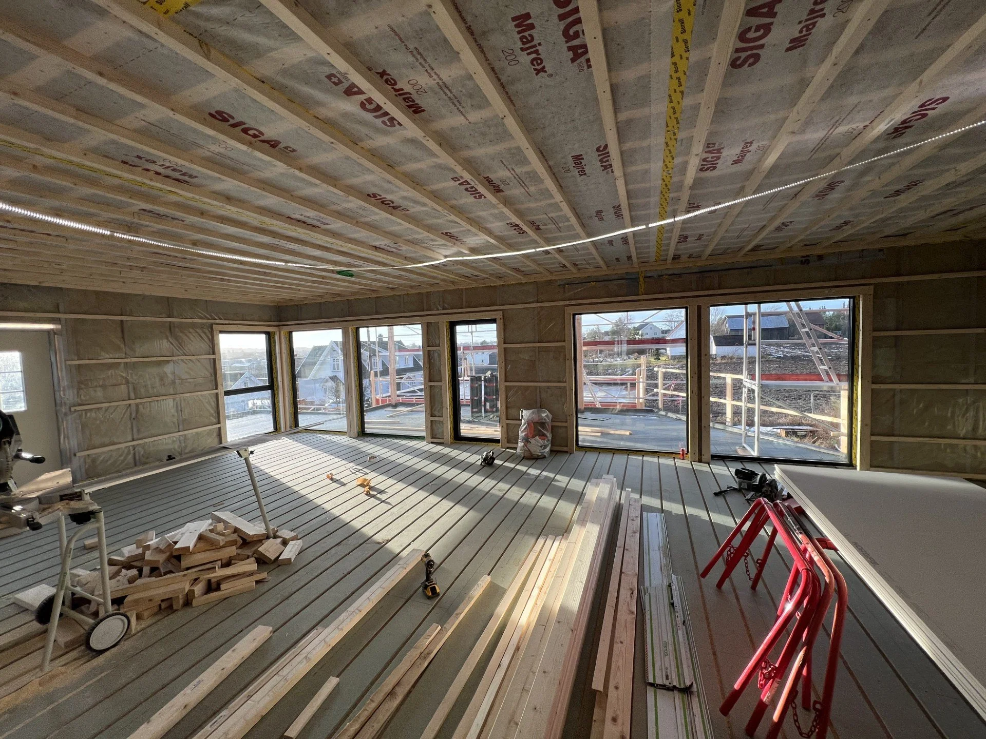 Interiør av et rom under bygging med store vinduer, uferdige vegger og gulv, og ulike byggeverktøy og materialer spredt rundt.