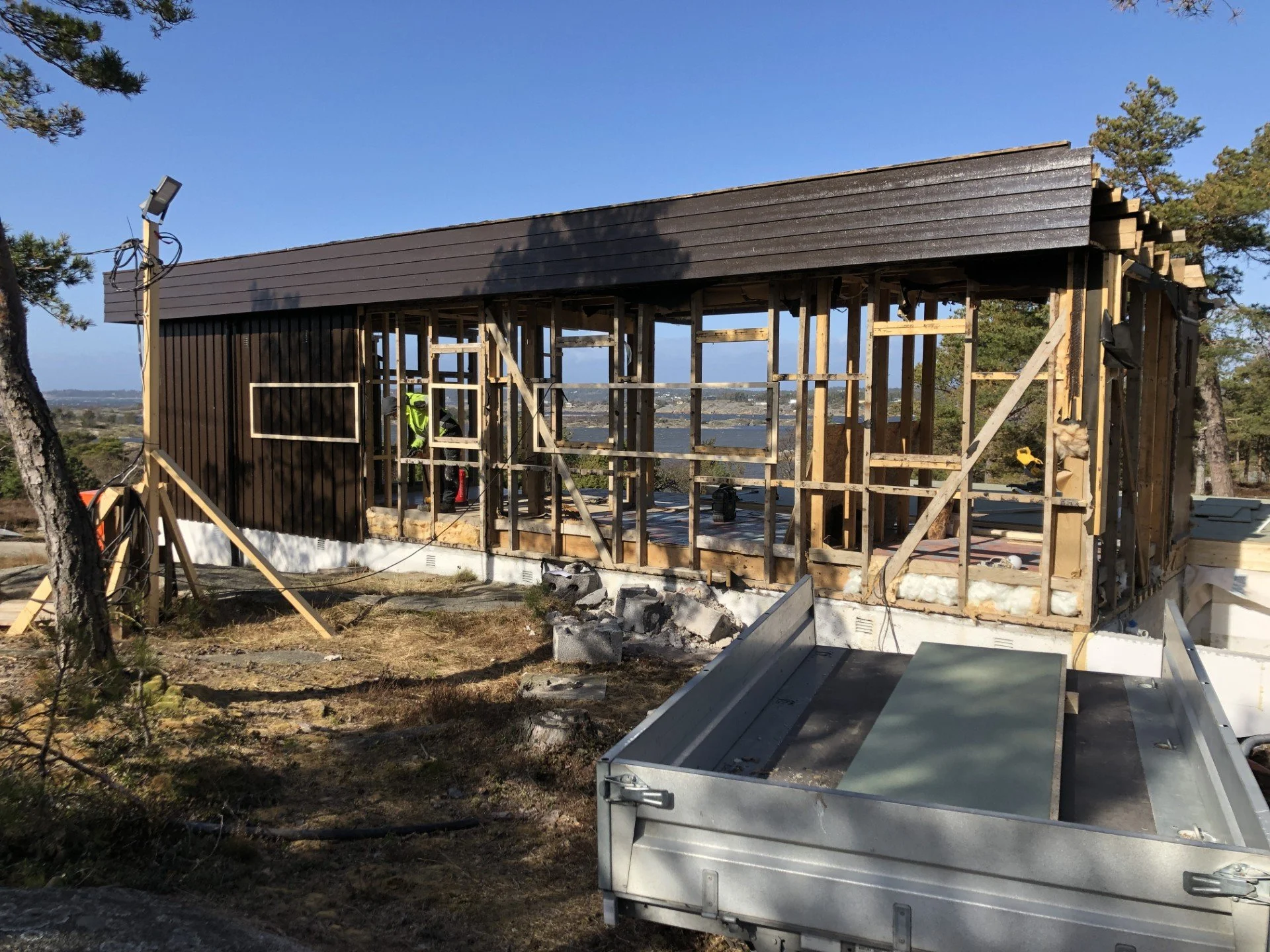Påbegynt trehus under konstruksjon med utvendig veggdekning delvis ferdig, omgitt av treverk og byggemateriell, med utsikt over sjøen i bakgrunnen.