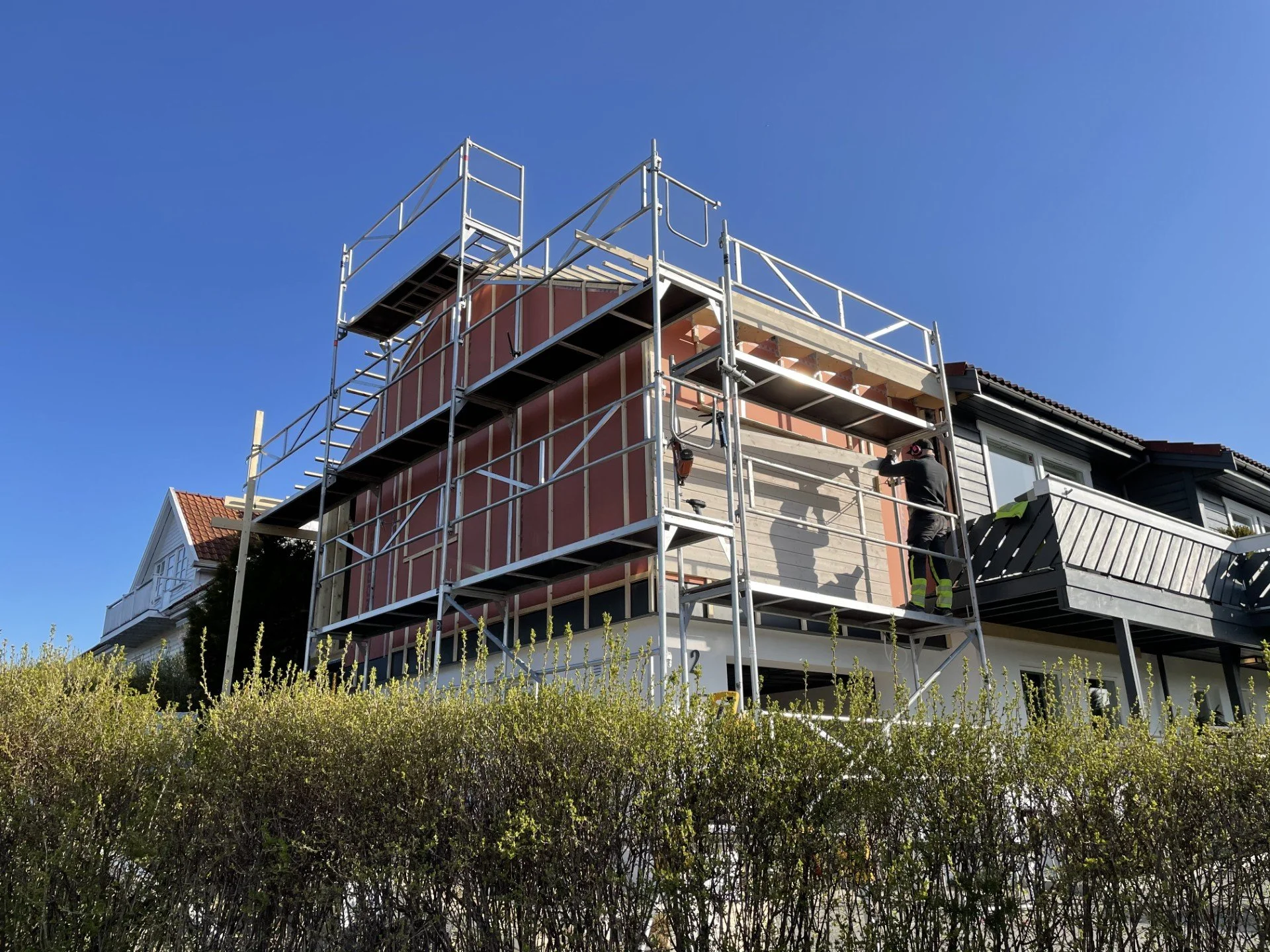 Et hus under bygging med scaffolding rundt, en mann som jobber på veggen, og en blå himmel.