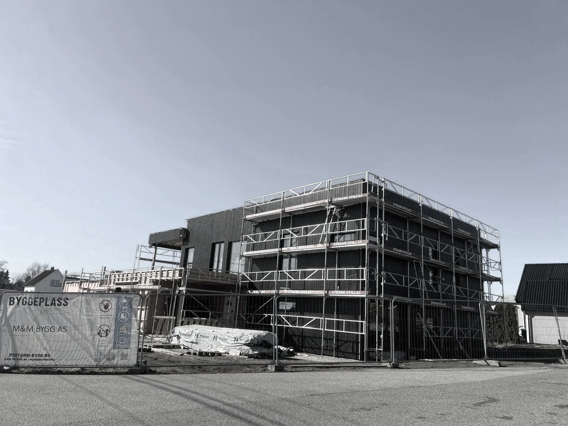 Byggeliv med flere etasjer under oppføring, omgitt av stillas, med skilt som sier BYGGEPLASS, i et boligområde med eksisterende boliger.